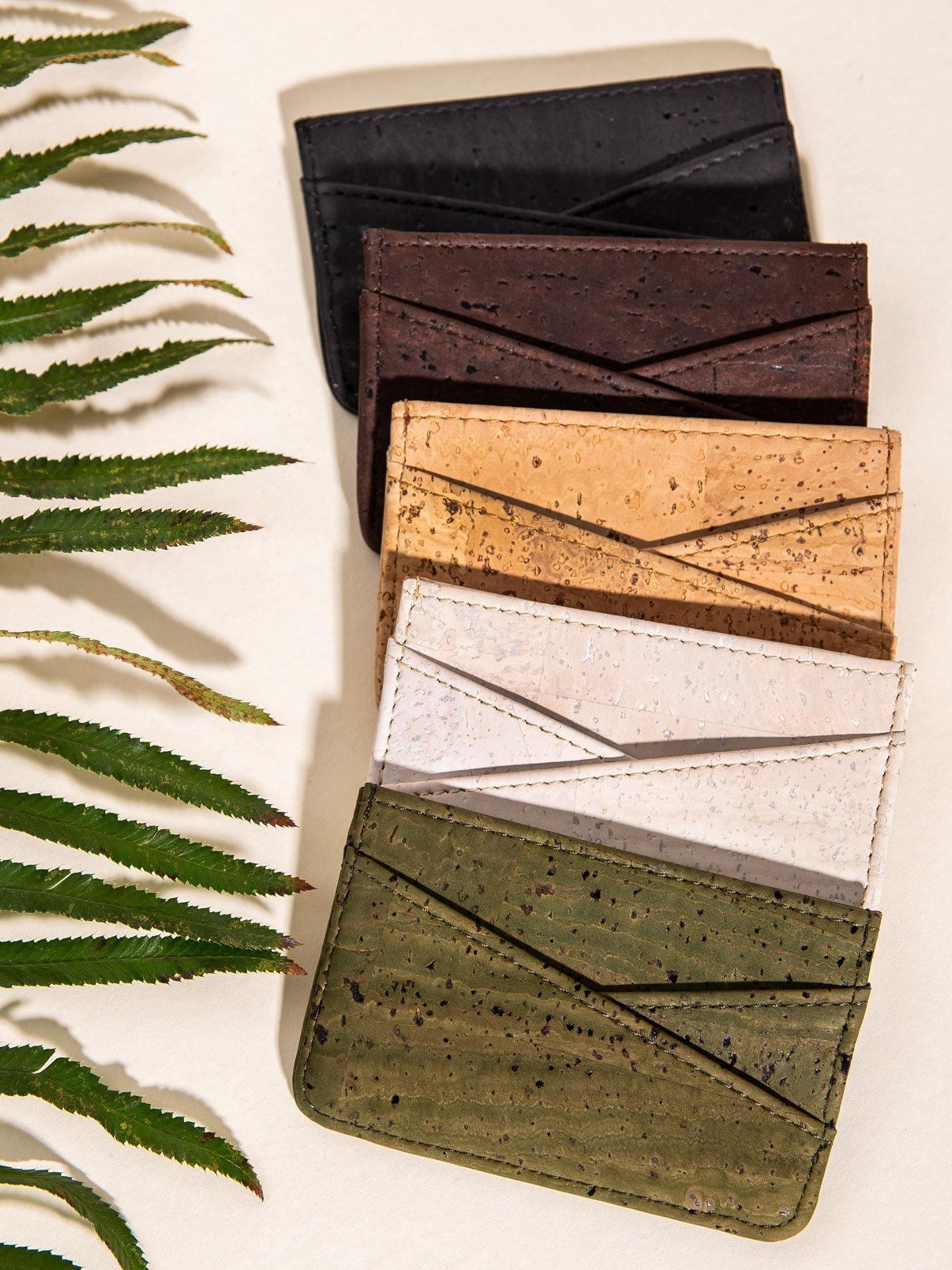 Five cork card wallets in black, brown, tan, white, and green on cream background with fern leaf