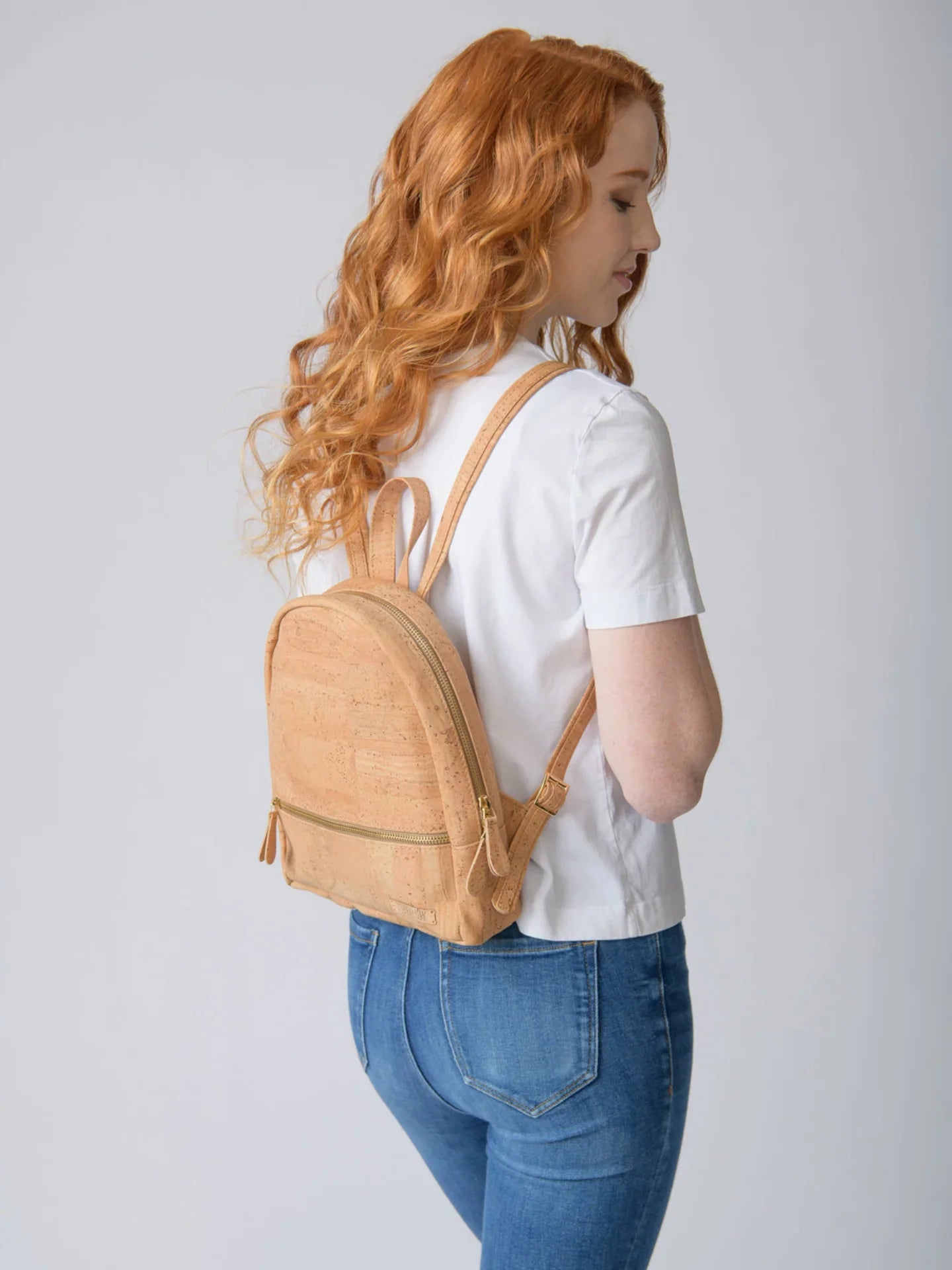 Woman with wavy red hair wearing white shirt, jeans, and tan cork backpack from Qu!rk Boutique