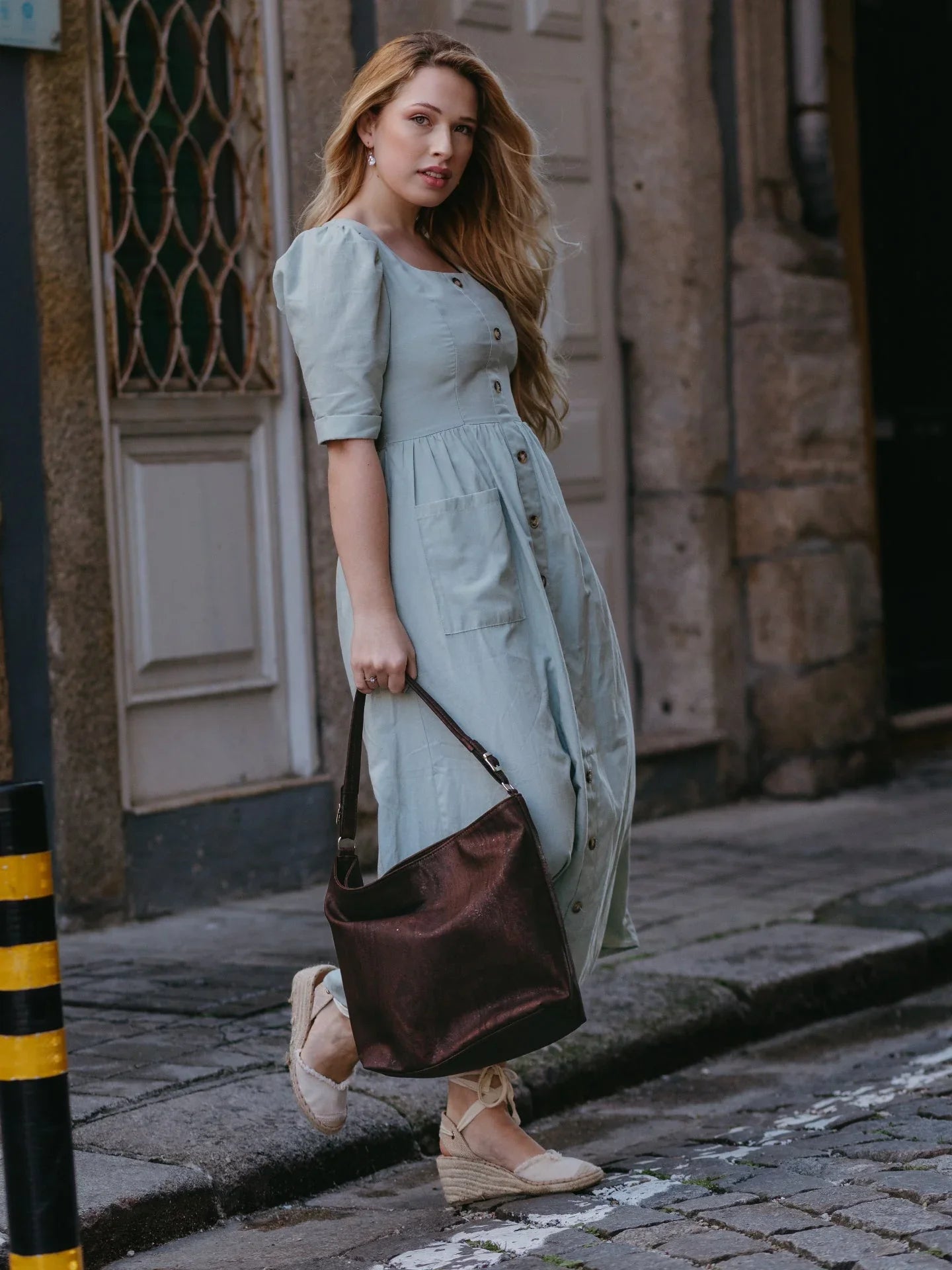 Woman in a light blue button-front dress and espadrilles holding a brown tote on a cobblestone street