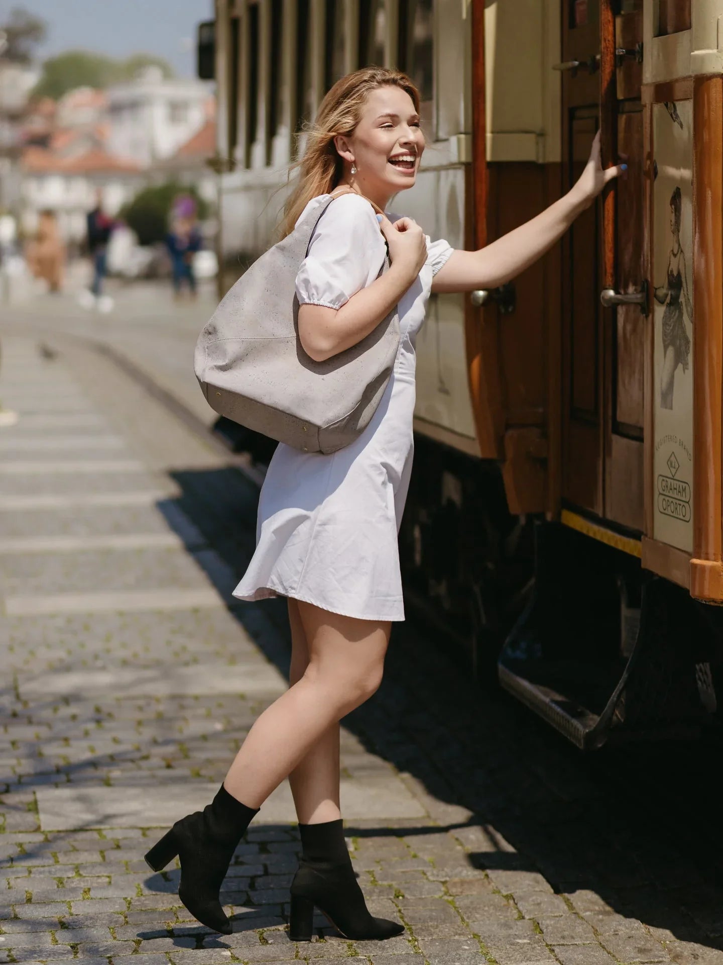 Woman in a white dress with black ankle boots and a large tote bag boarding a vintage tram outdoors.