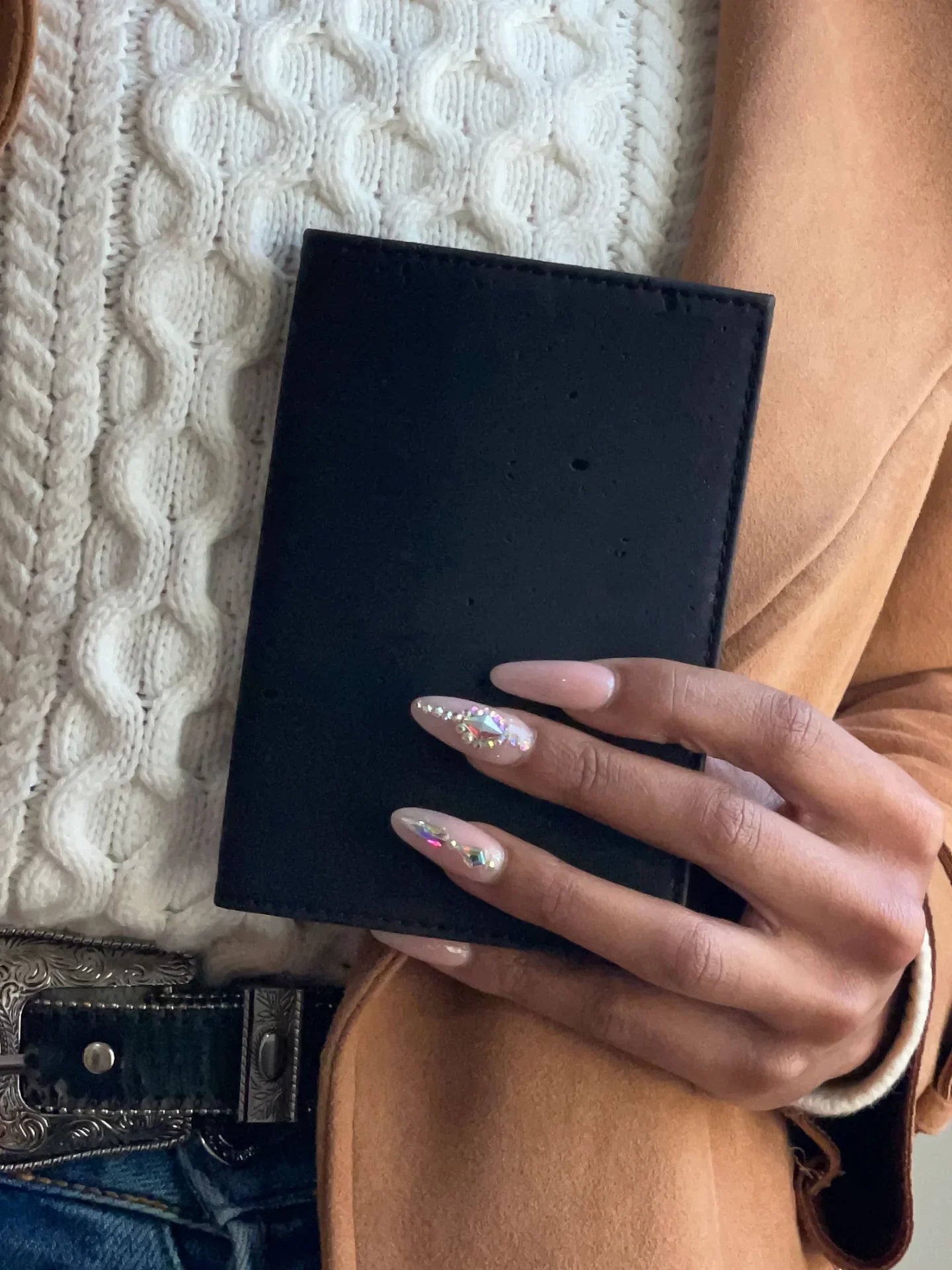 Woman in cozy sweater and tan coat holding a black wallet, featuring jeweled nails
