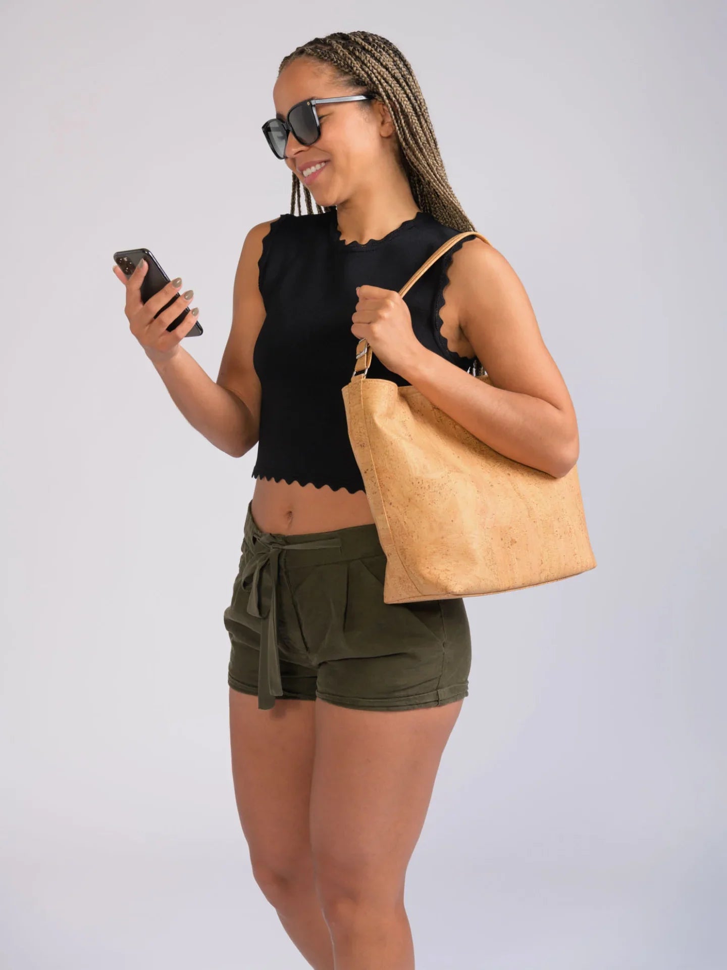 Woman wearing black crop top, olive shorts, and sunglasses, holding cork handbag and phone