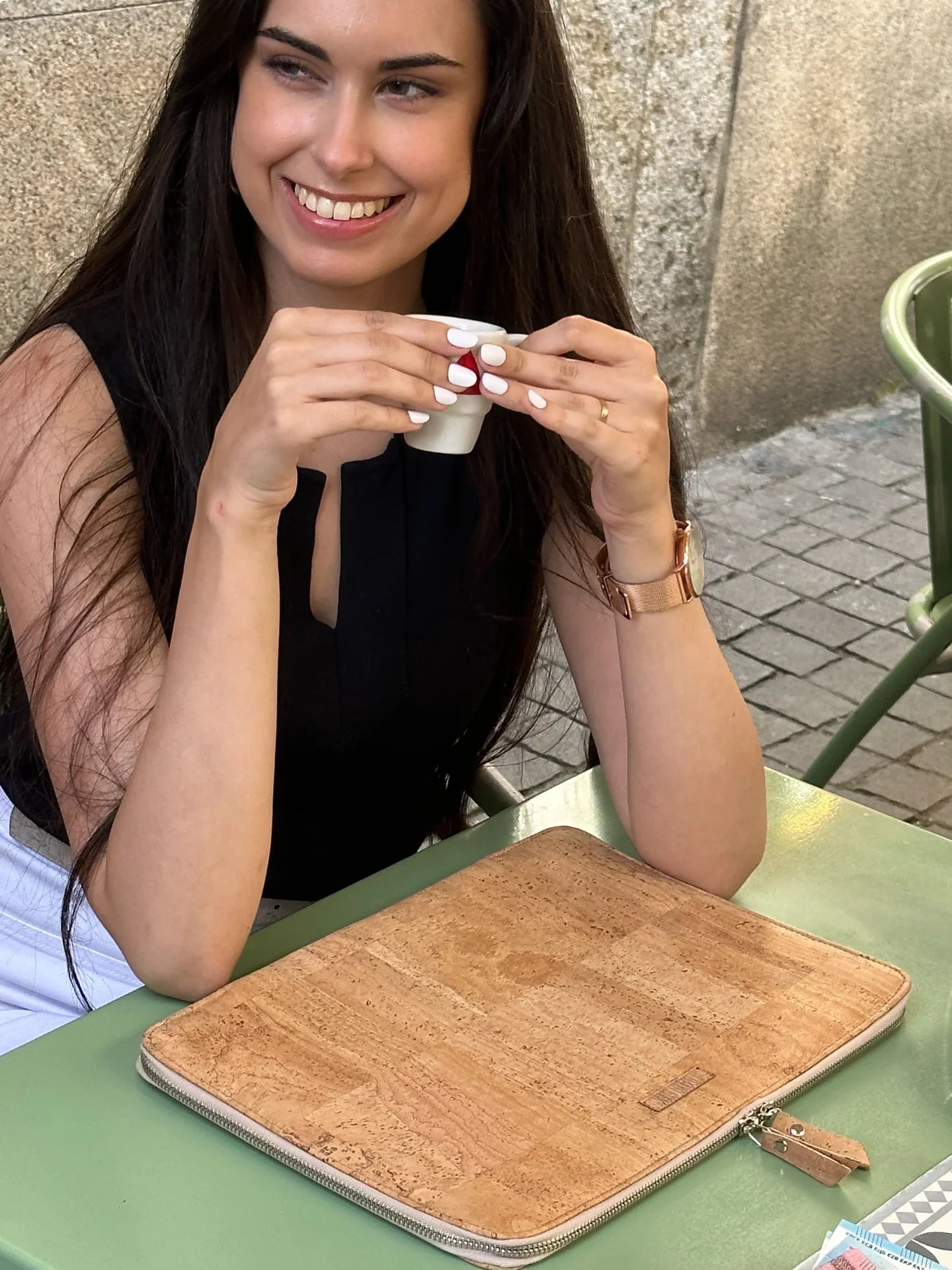Woman smiling outdoors at café table with natural cork laptop sleeve, sustainable fashion accessory
