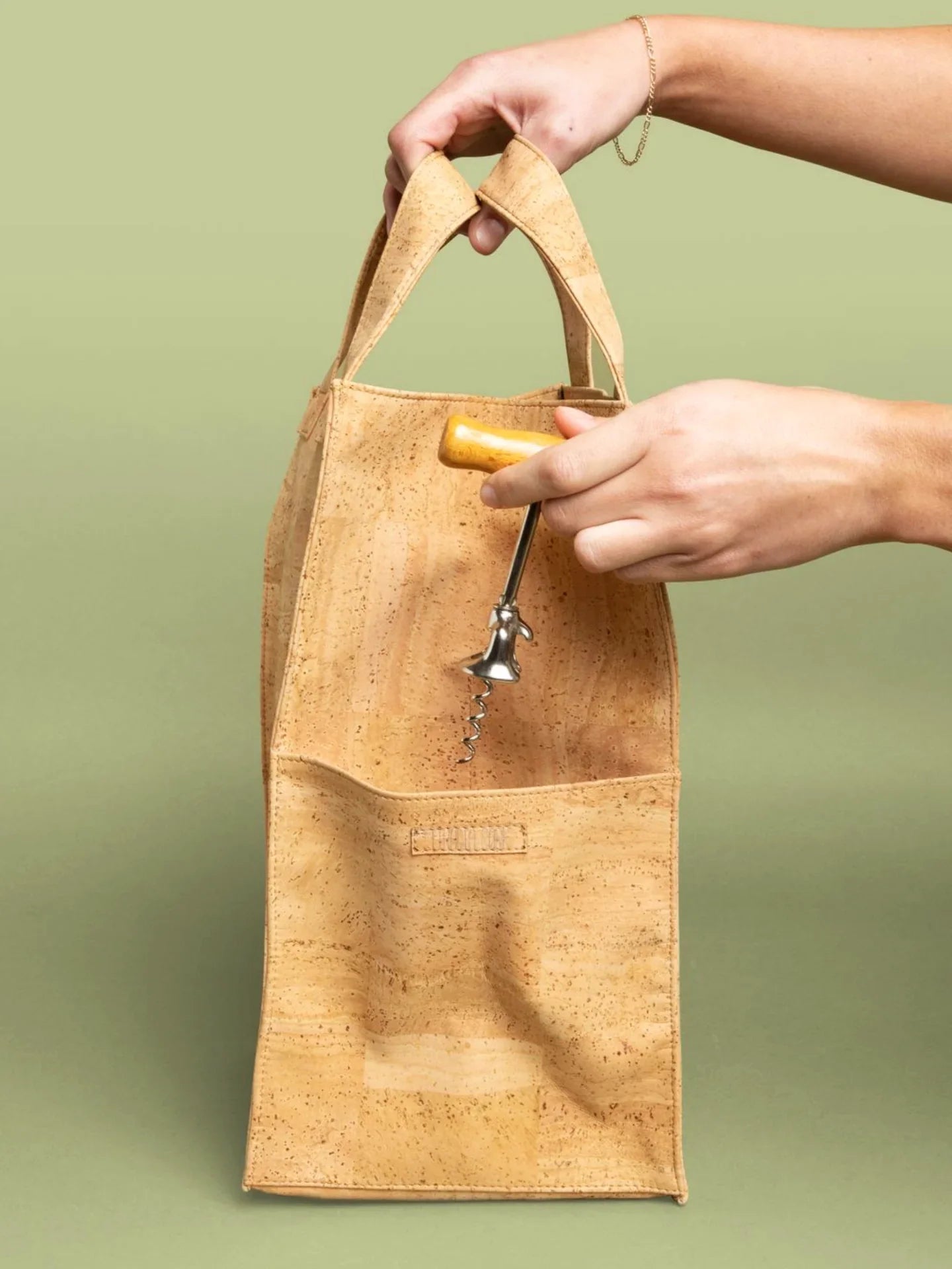 Hand placing corkscrew into cork wine tote bag on green background, Qu!rk Boutique gift