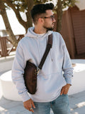 Man in light hoodie and sunglasses with brown crossbody bag, outdoor winter boutique style