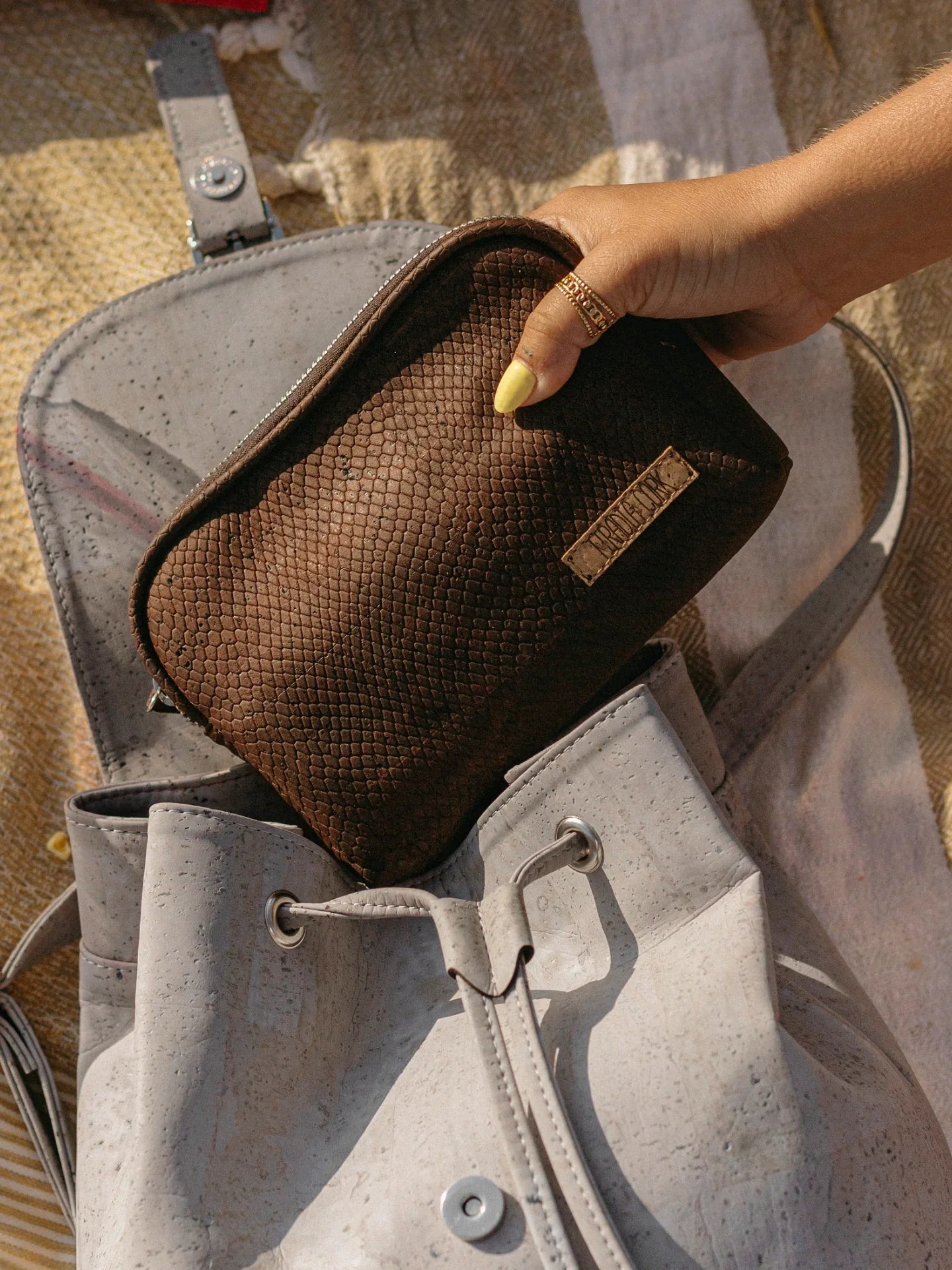 Hand with yellow nails holds textured brown pouch above light gray cork backpack on picnic blanket