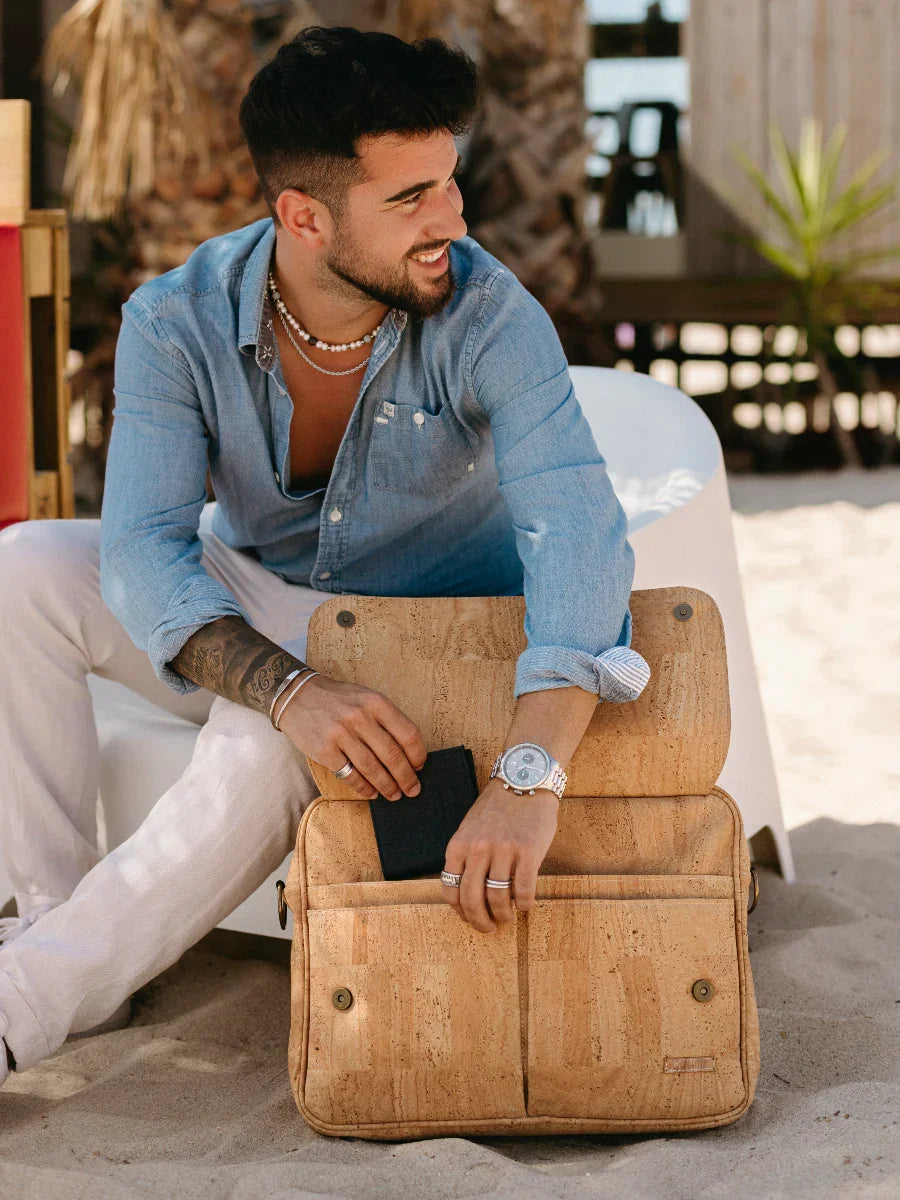 Man on sandy beach in blue shirt holding cork laptop bag and wallet, Qu!rk Boutique style