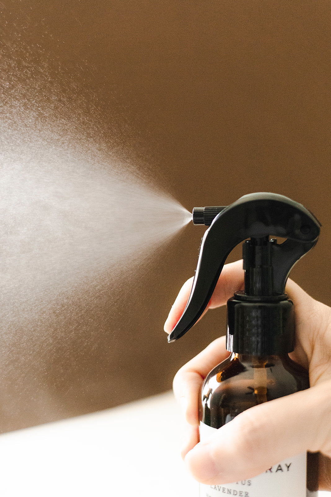 Close-up of hand spraying mist from amber glass spray bottle with black trigger on brown background
