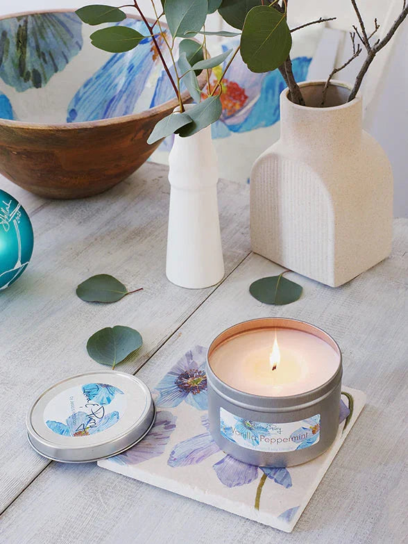 Vanilla peppermint scented candle in metal tin on floral cloth with white ceramic vases and eucalyptus leaves