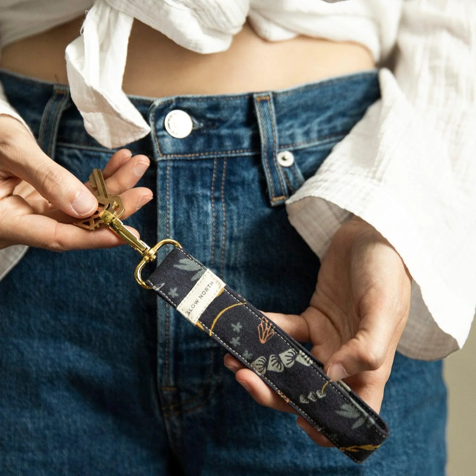 Person holding a slow north branded floral fabric wristlet keychain attached to gold keys, wearing blue jeans and white shirt
