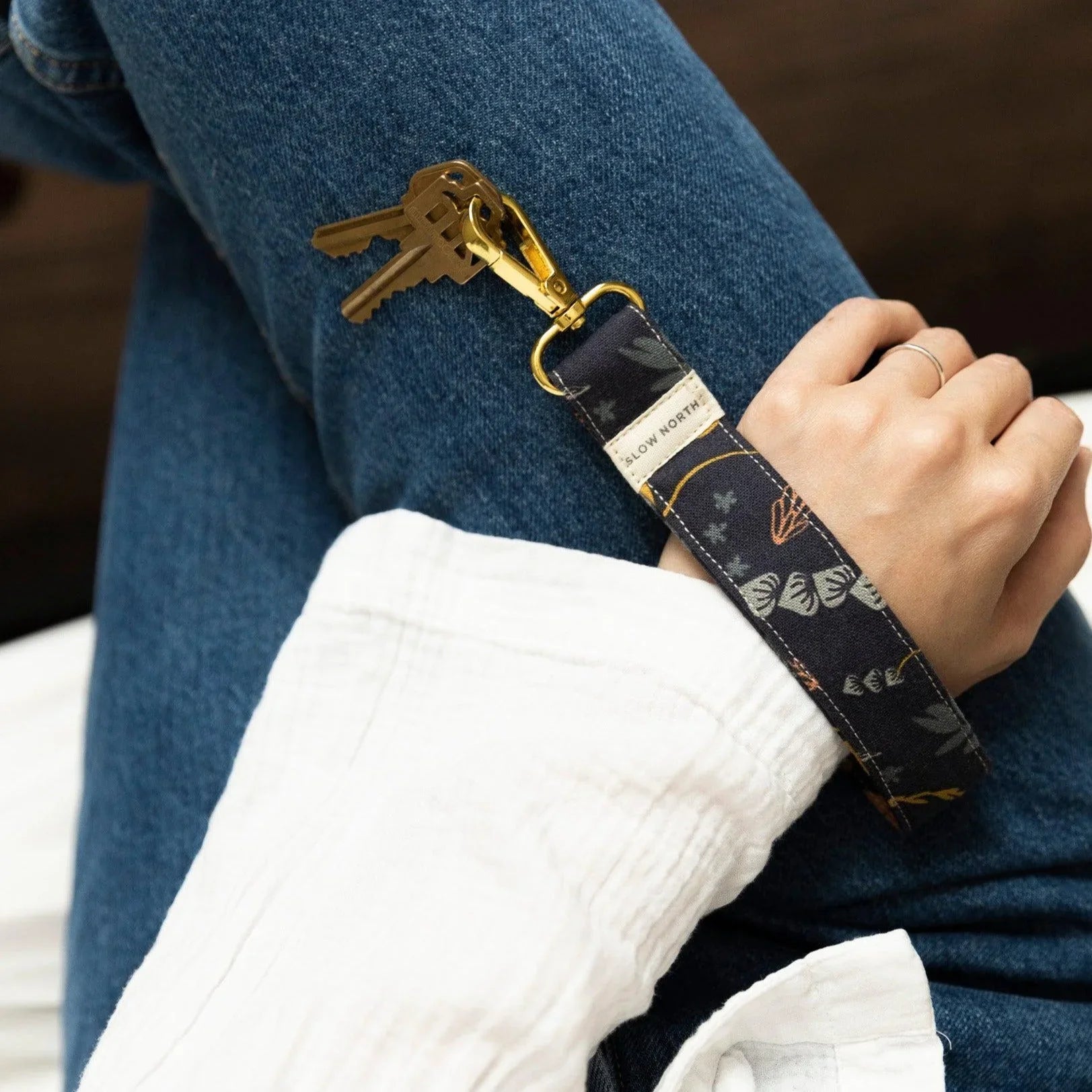 Person wearing blue jeans and white sleeve holding keys with a black floral Slow North wristlet keychain