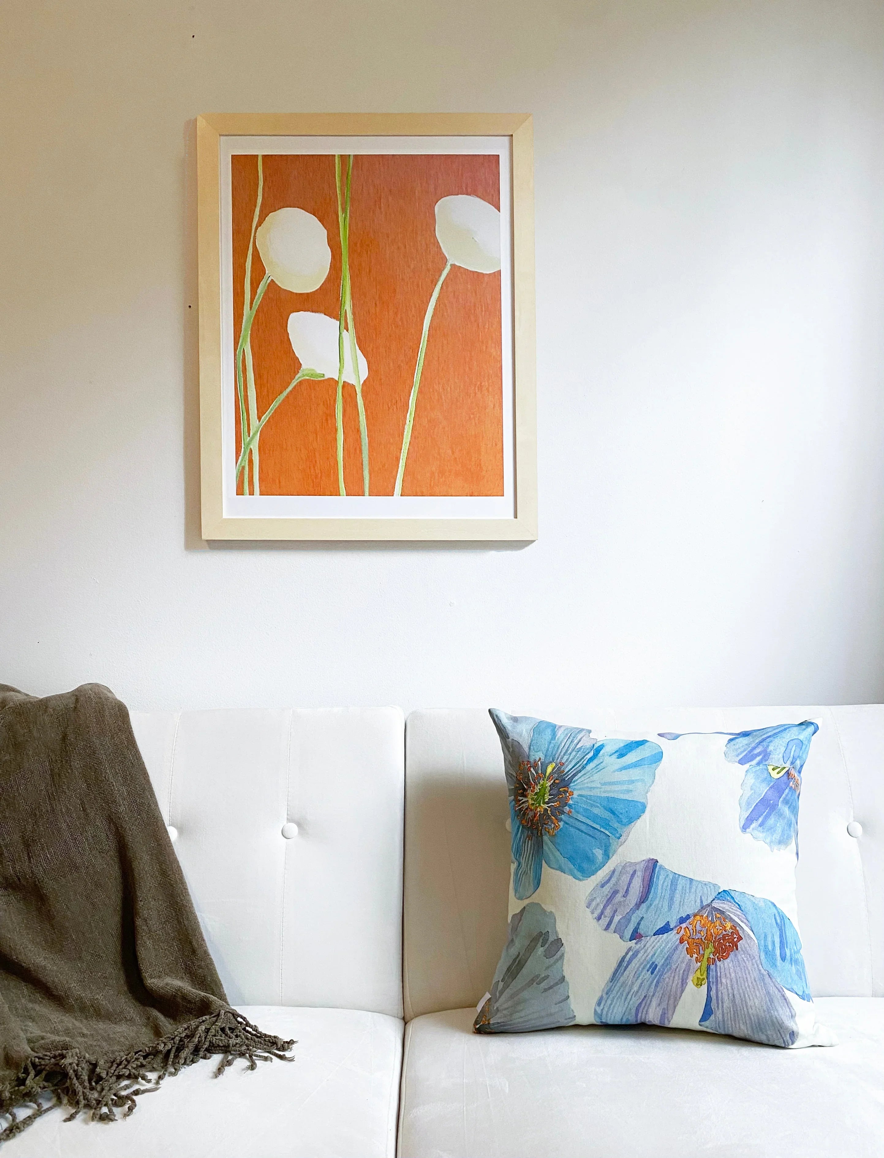 White couch with blue floral pillow and brown throw, orange floral art framed on white wall