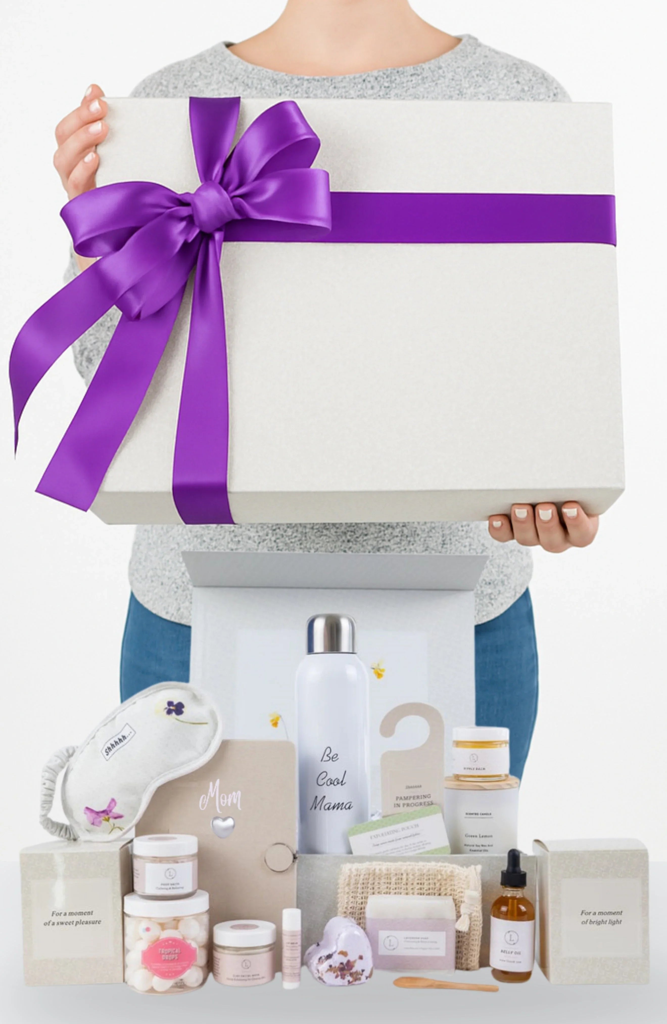 Person holding large white gift box with purple ribbon above a new mom self-care gift set
