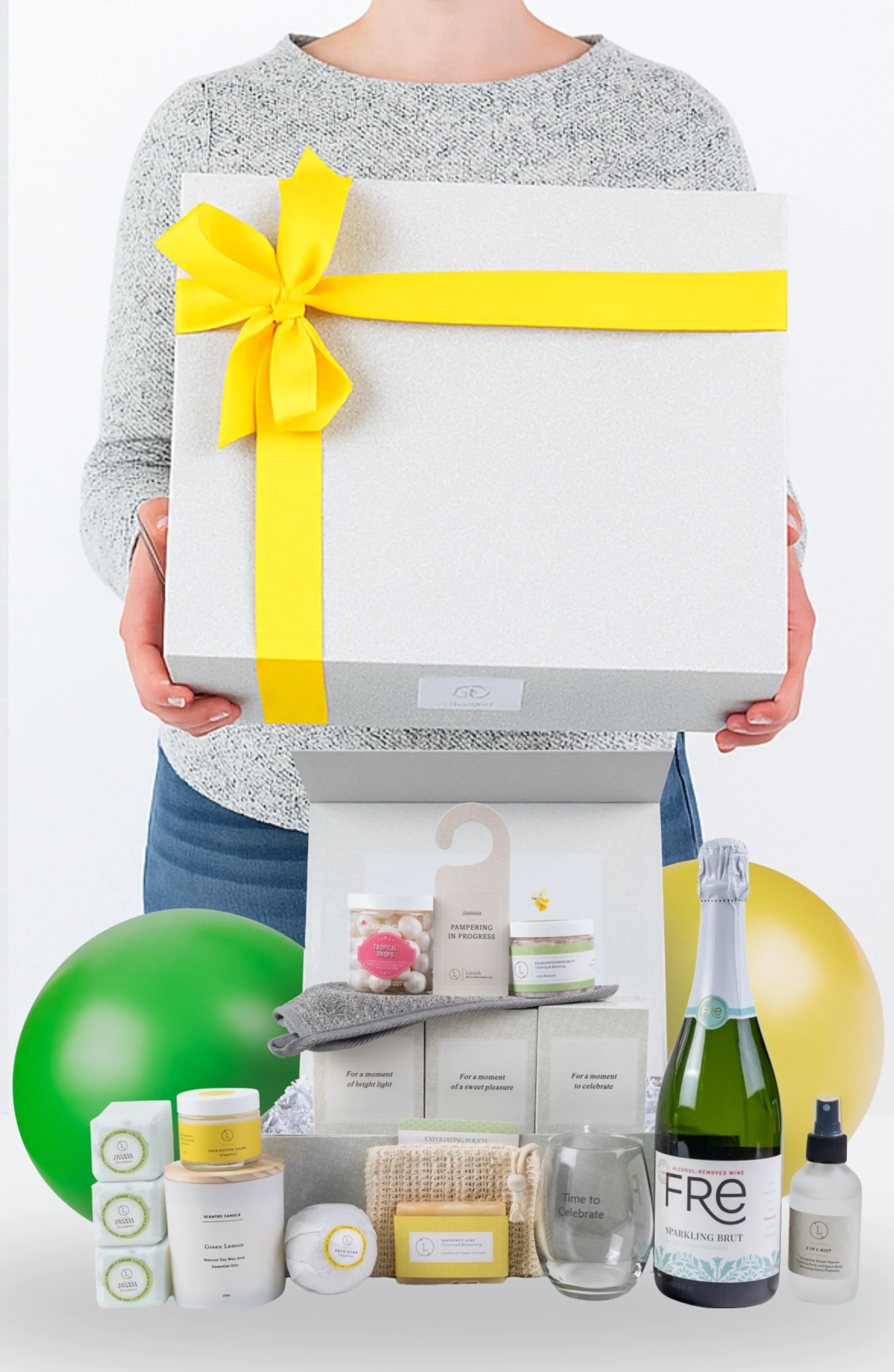 Person holding large gift box with yellow ribbon and assorted spa and celebration products inside
