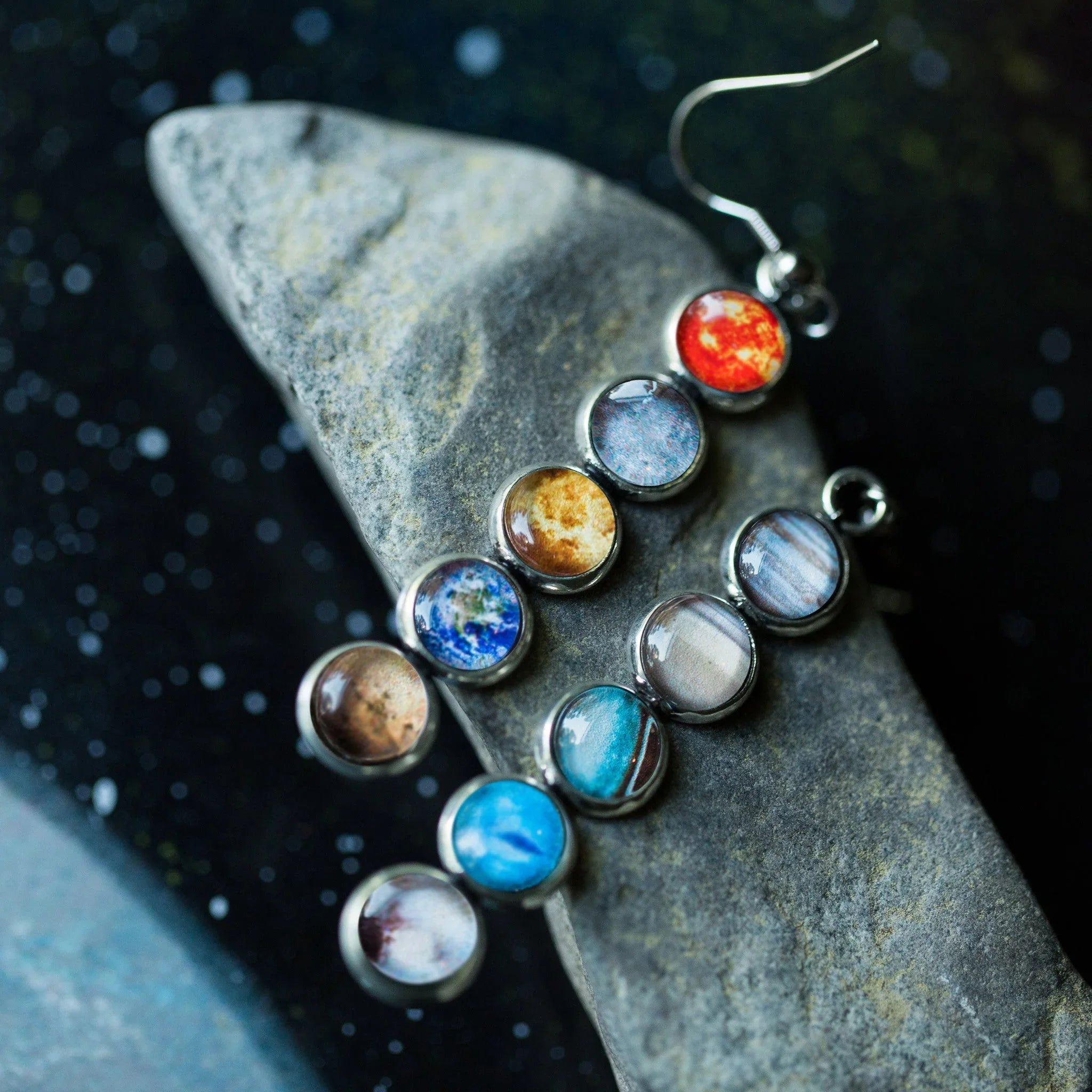 Silver earrings featuring colorful planets with space background on stone surface