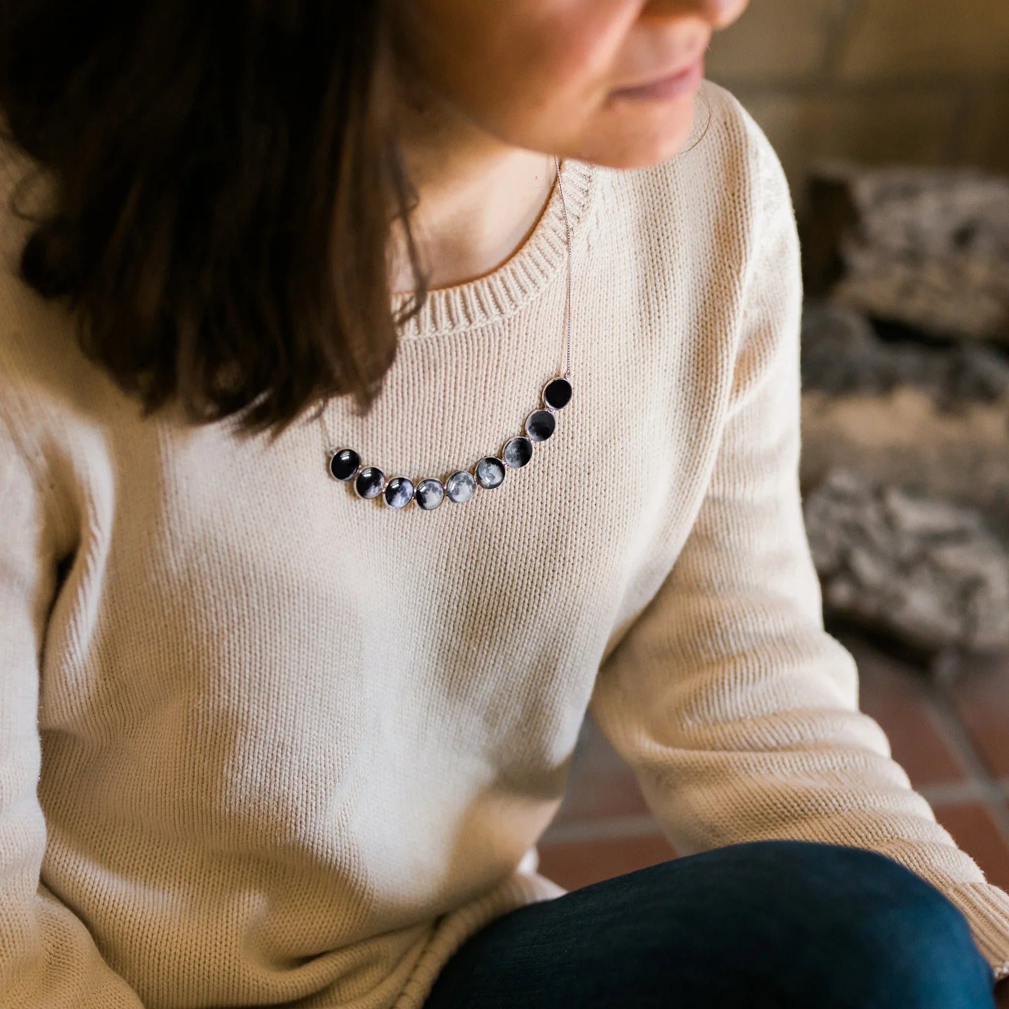 Woman wearing beige sweater and moon phase necklace sitting indoors with stone fireplace background