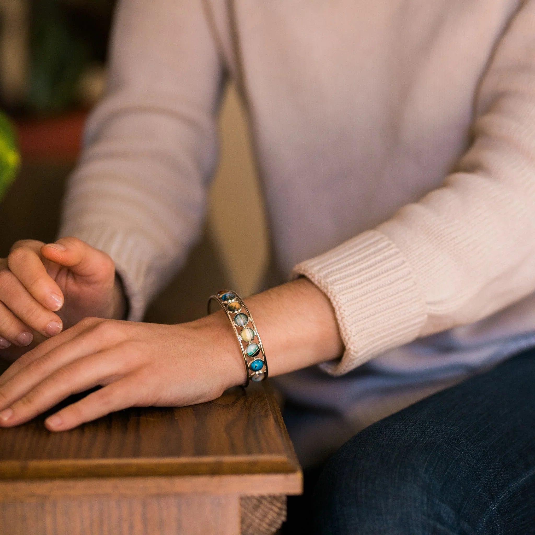 Close-up of a person wearing a colorful stone bracelet and a beige sweater sitting at a wooden table