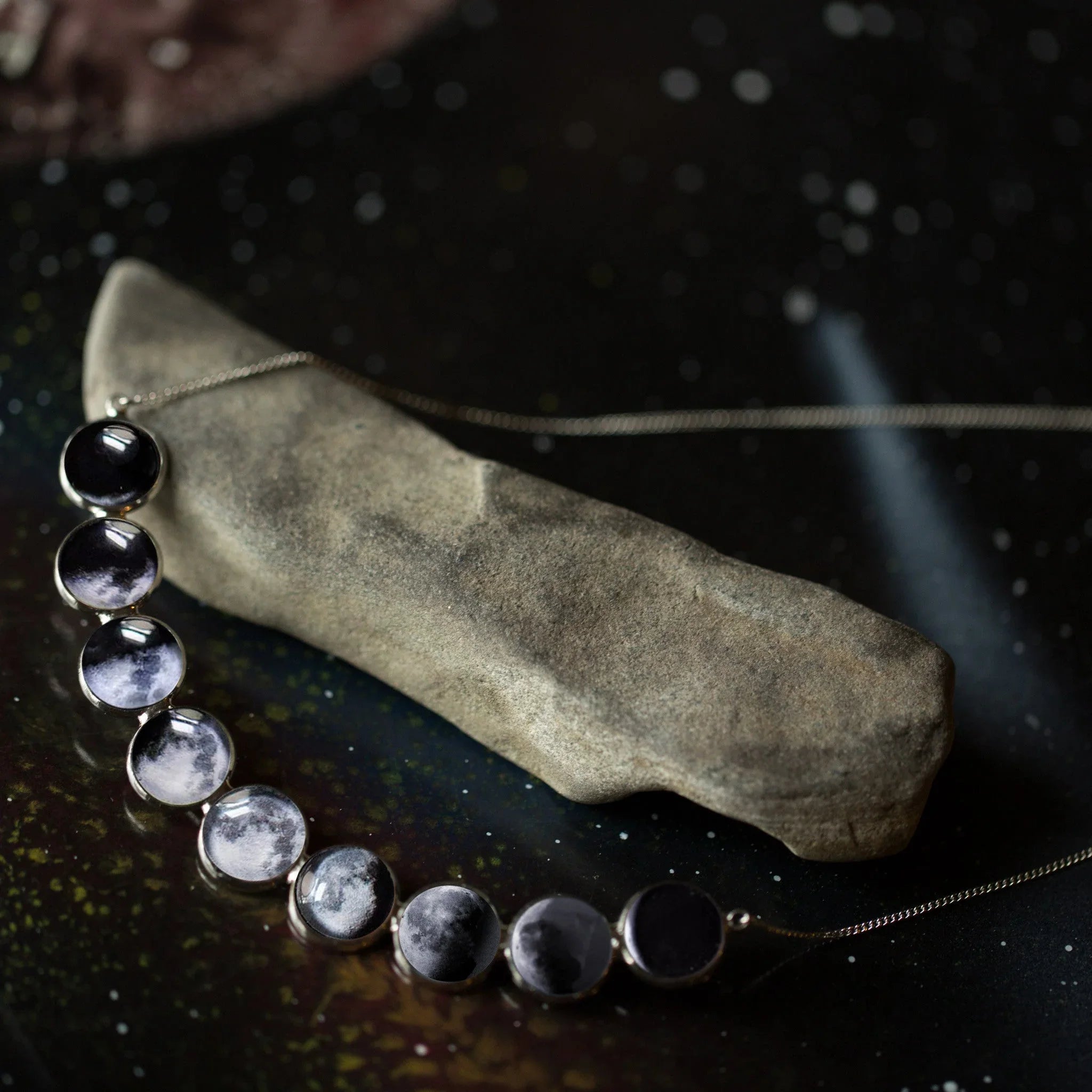 Close-up of a silver moon phase necklace displayed on an irregular stone against a dark starry background