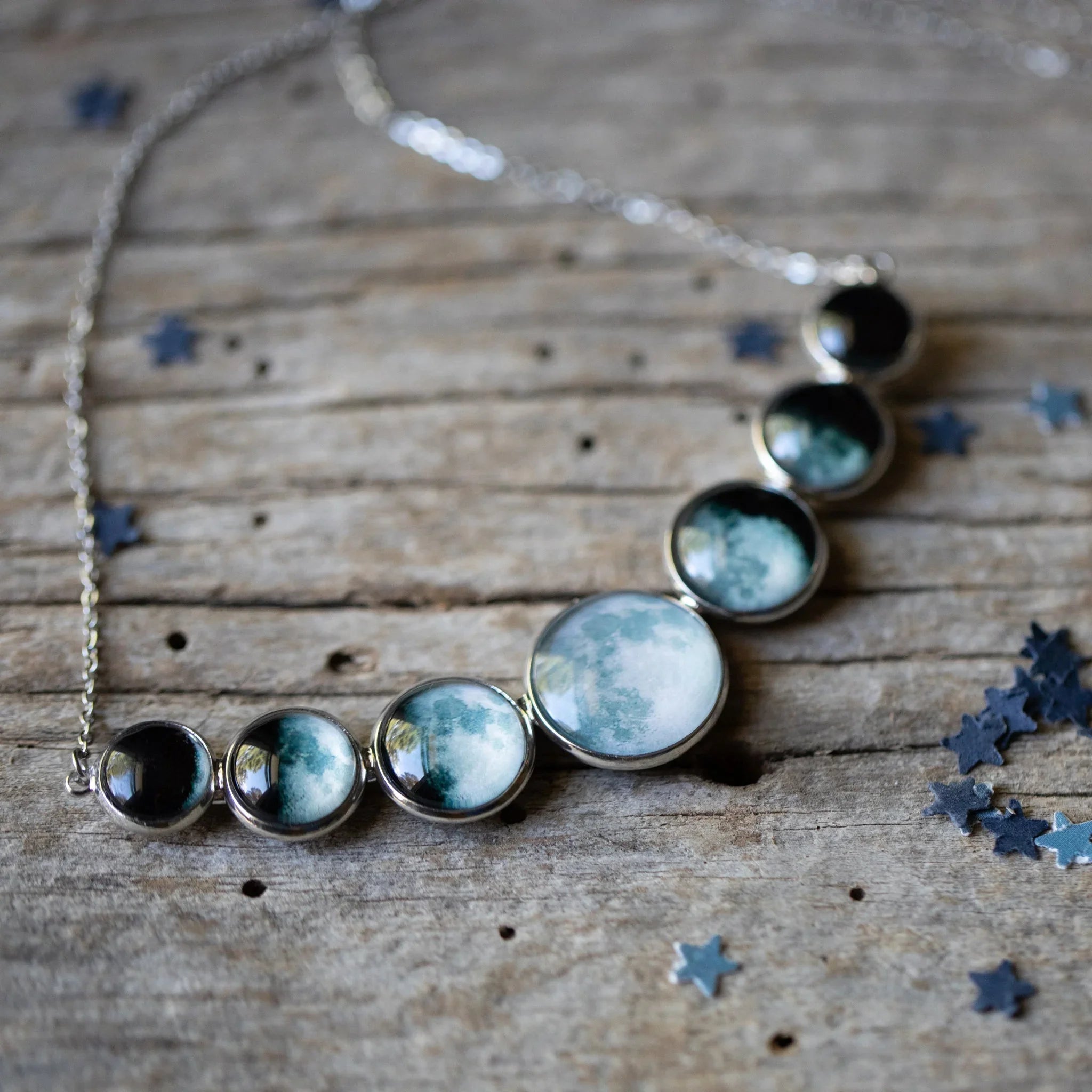 Silver necklace with seven round moon phase pendants on rustic wooden surface with star confetti