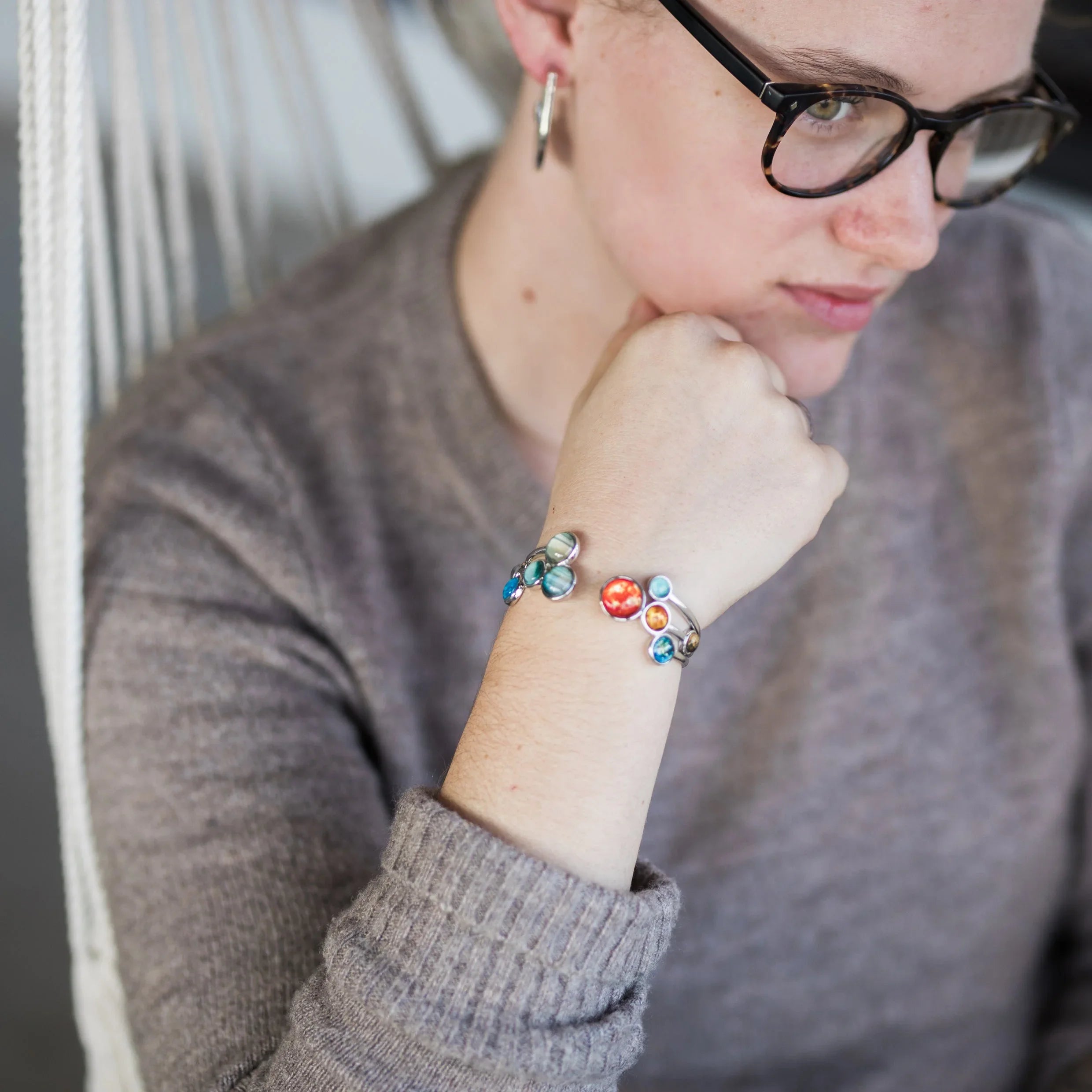 Woman wearing glasses and gray sweater displaying silver bracelet with colorful planet charms