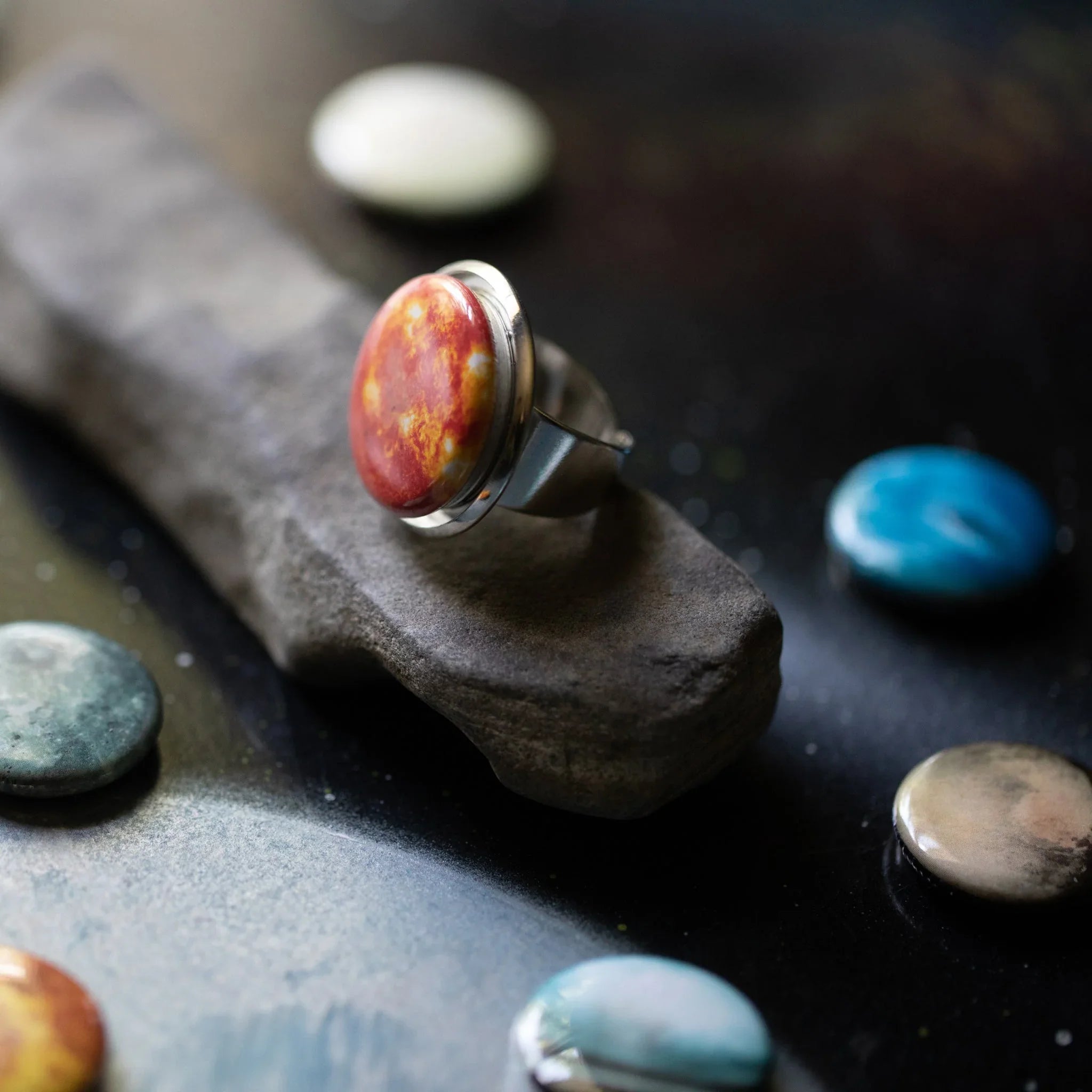 Close-up of a silver ring with a round red and orange gemstone on a textured stone display with colorful flat stones nearby