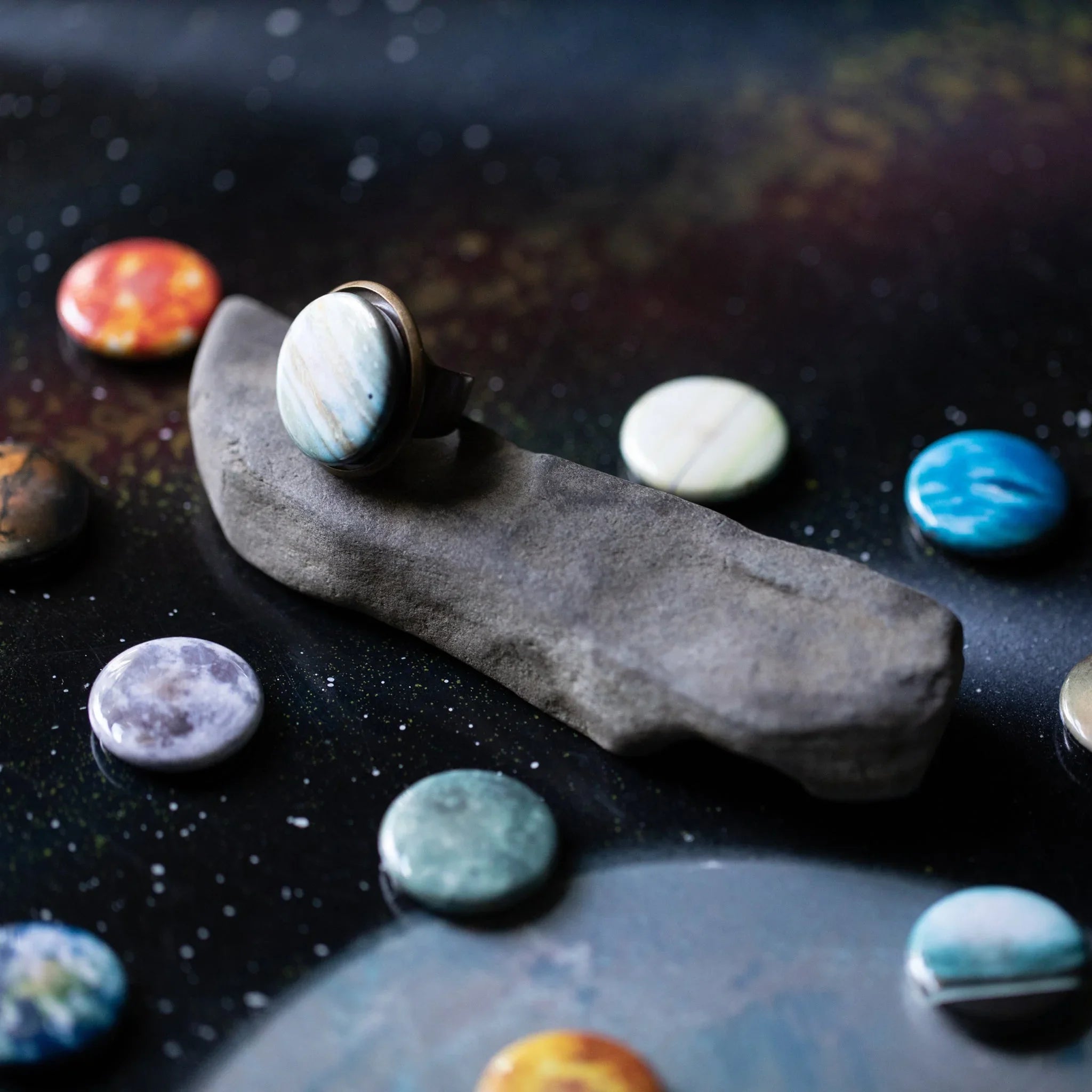 Ring with a round light blue stone on a gray rock surrounded by colorful circular stones on a dark speckled surface