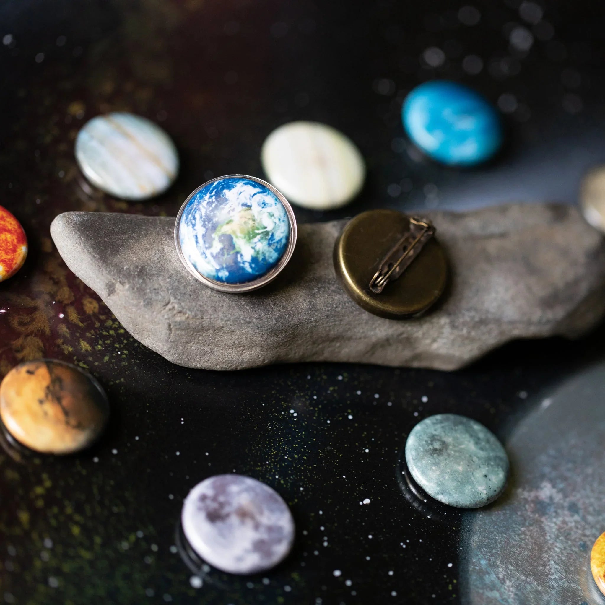 Round planet-themed pins, including Earth design, displayed on a stone and dark speckled surface