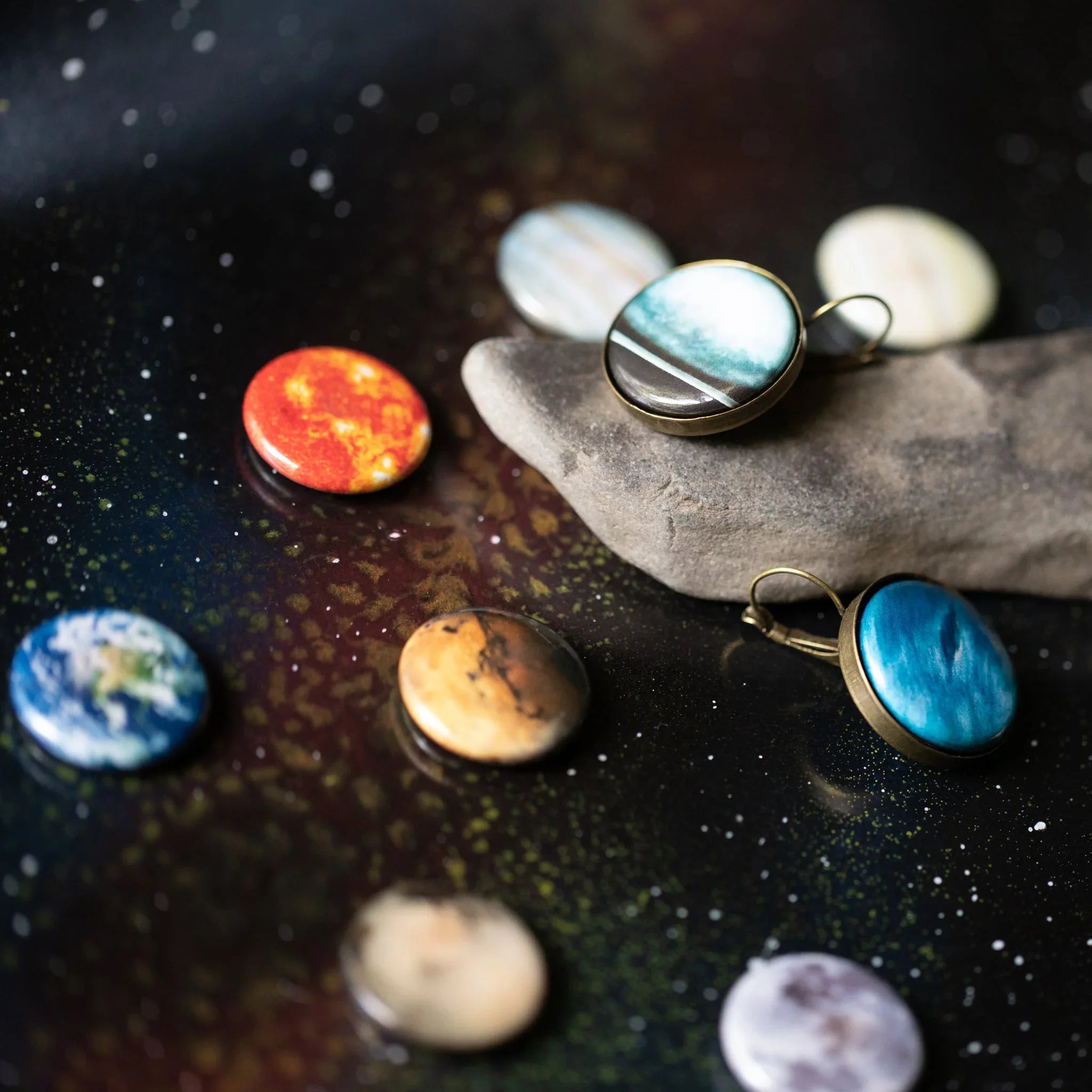 Round planetary-themed buttons and earrings displayed on a space-inspired background with a piece of driftwood