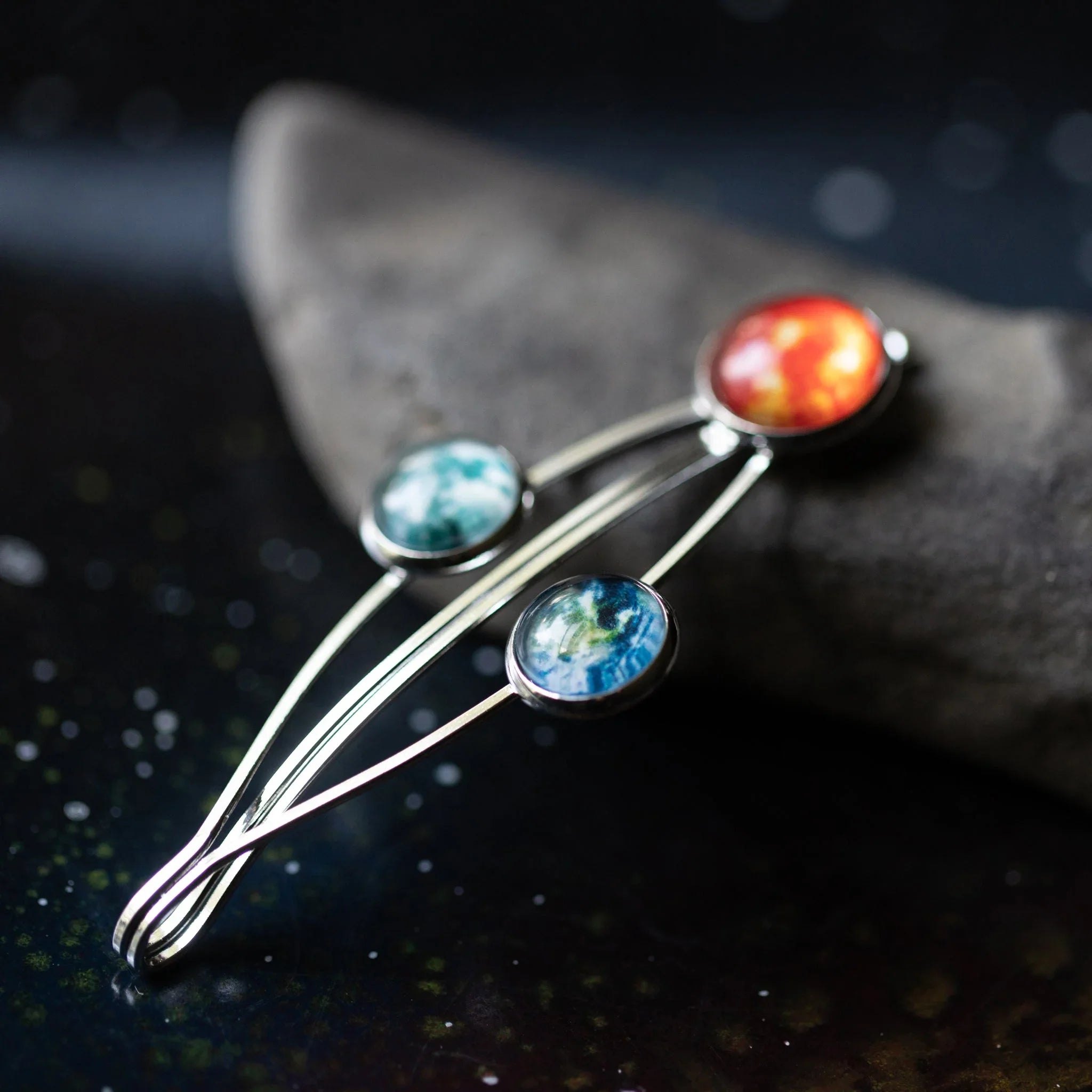 Silver hairpin with red, blue, and turquoise planet-like glass cabochons on dark textured surface