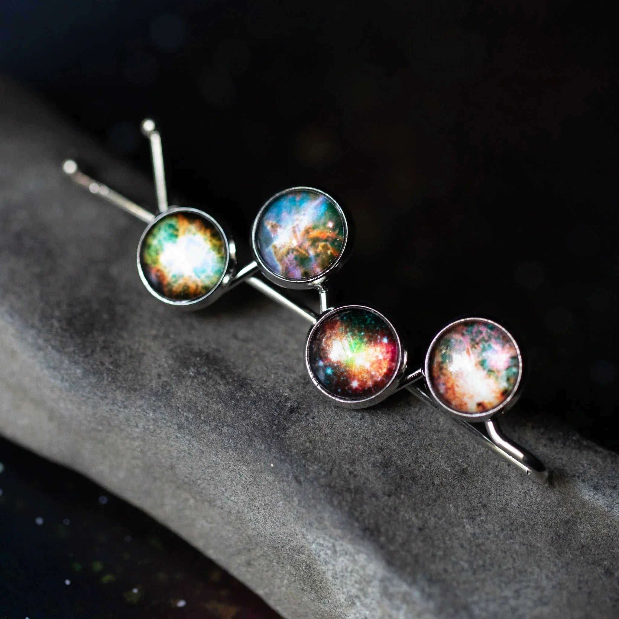 Cosmic galaxy hair pins with colorful nebula designs, displayed on a stone, Qu!rk Boutique