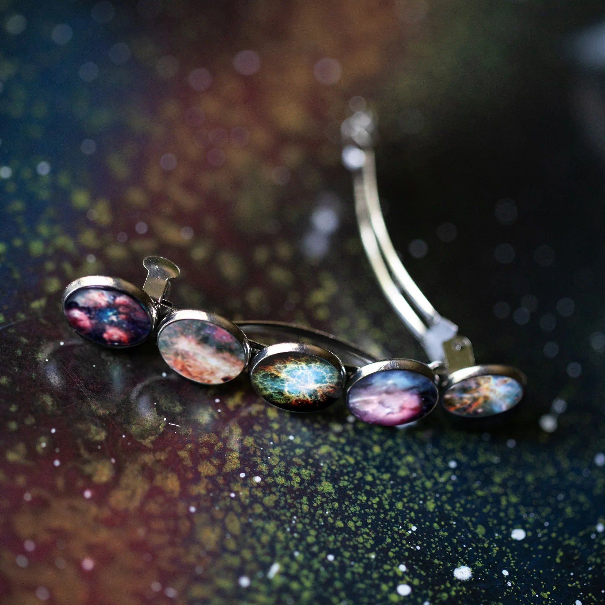Silver hair clip with five oval galaxy and nebula space designs on a speckled dark surface
