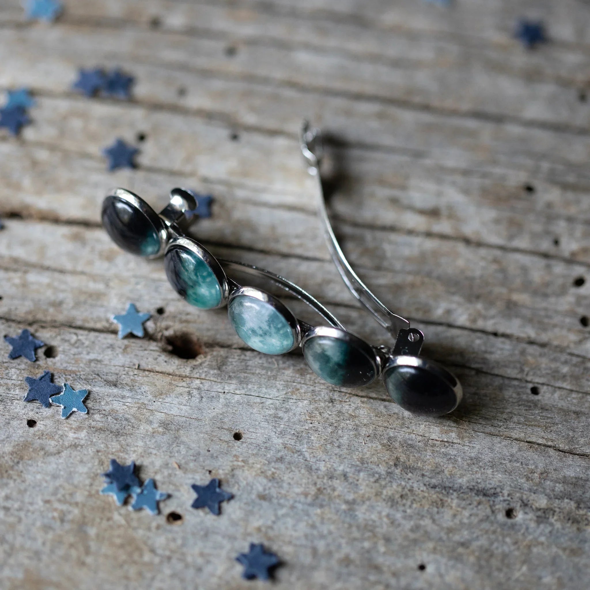 Silver hair clip with five round blue-green stones on rustic wooden surface with scattered blue star confetti
