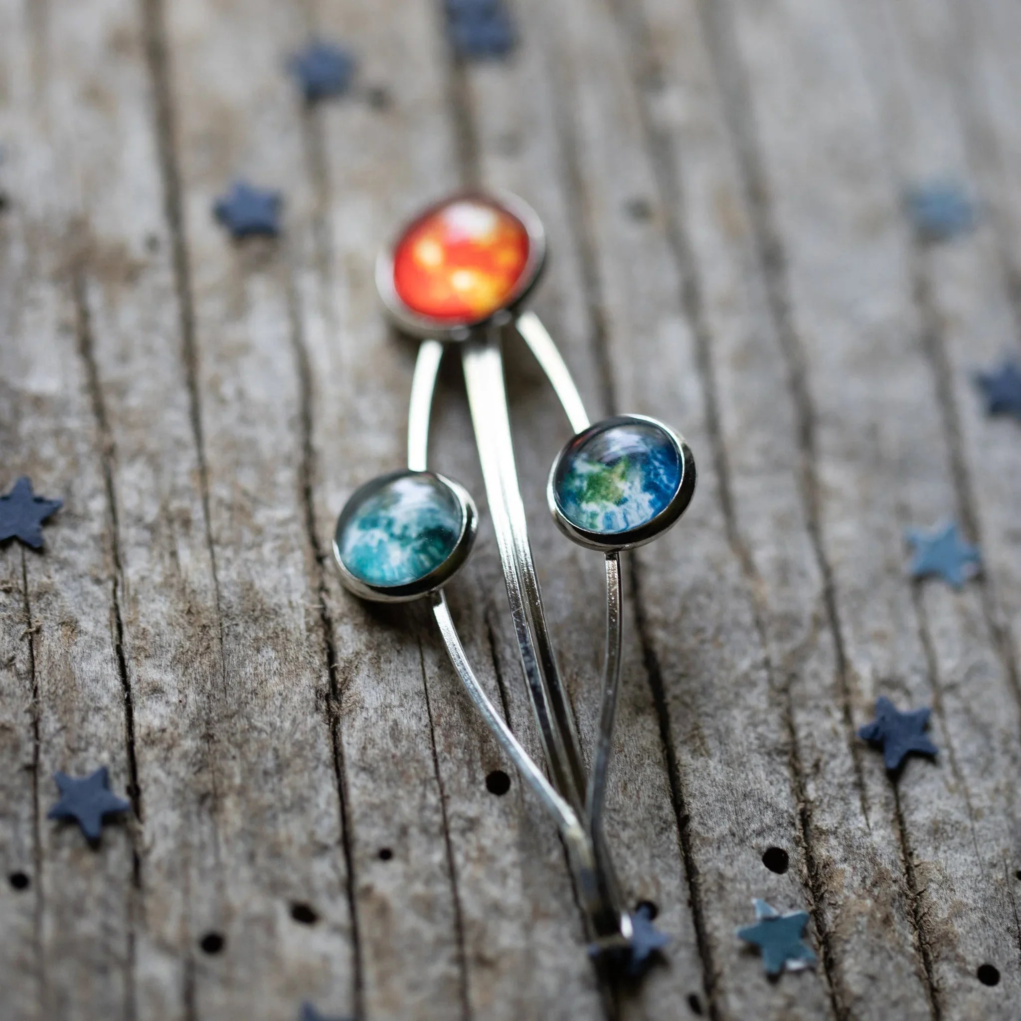 Celestial-themed silver brooch with red and blue enamel planets on rustic wooden background with star confetti