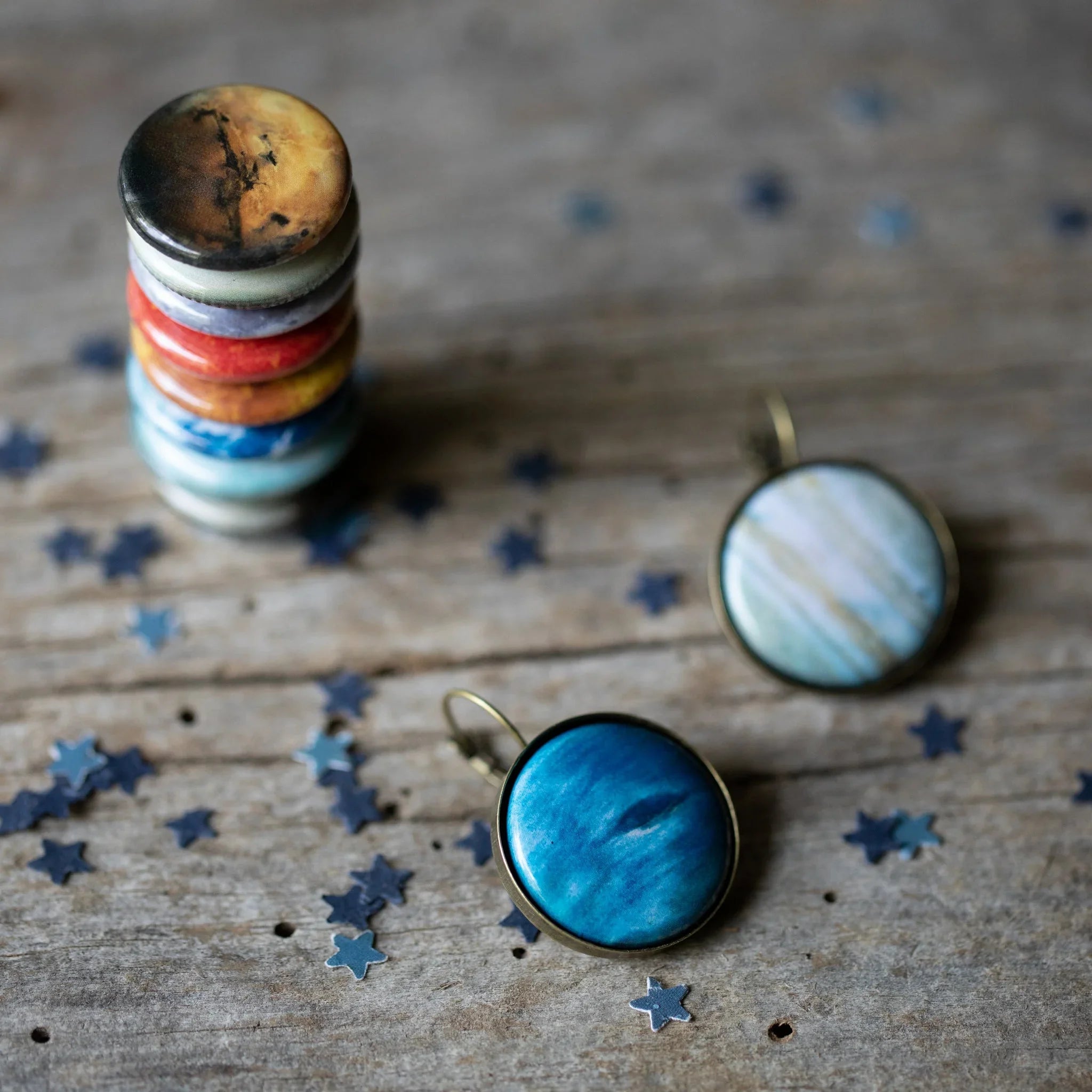 Stack of colorful round planet-themed pins and two planet print earrings on wooden surface with star confetti