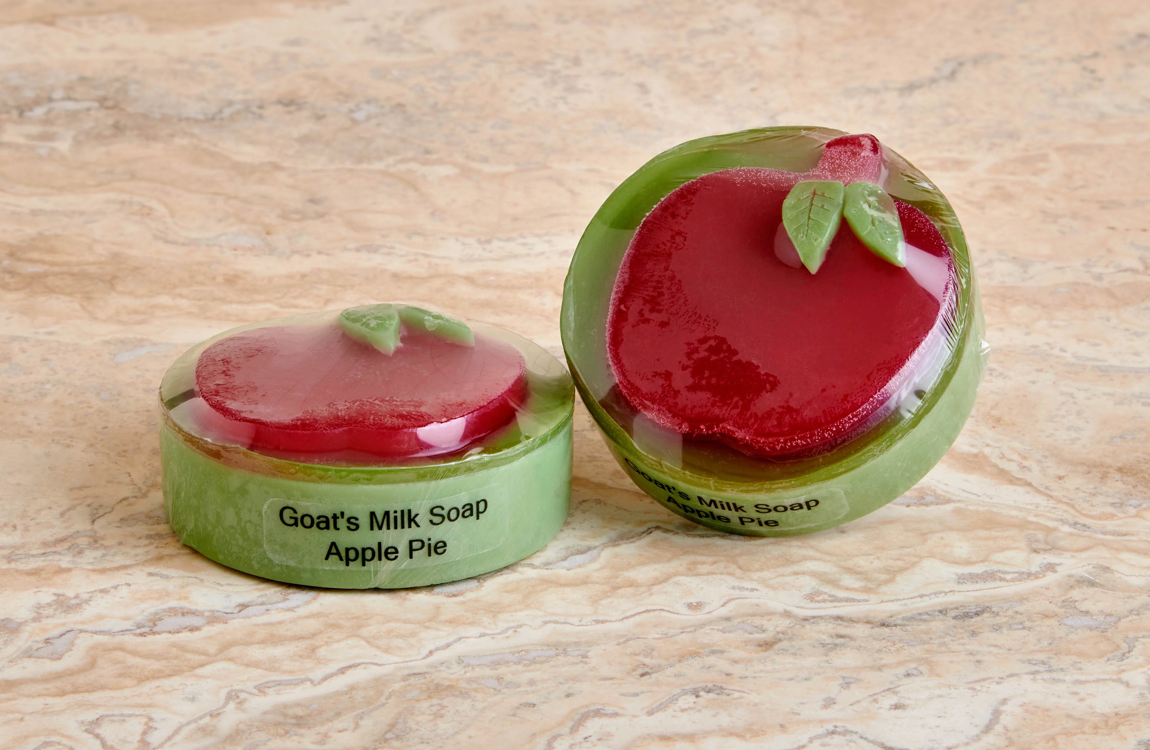 Two round goat's milk soap bars labeled Apple Pie with red apple-shaped tops and green bases on marble surface