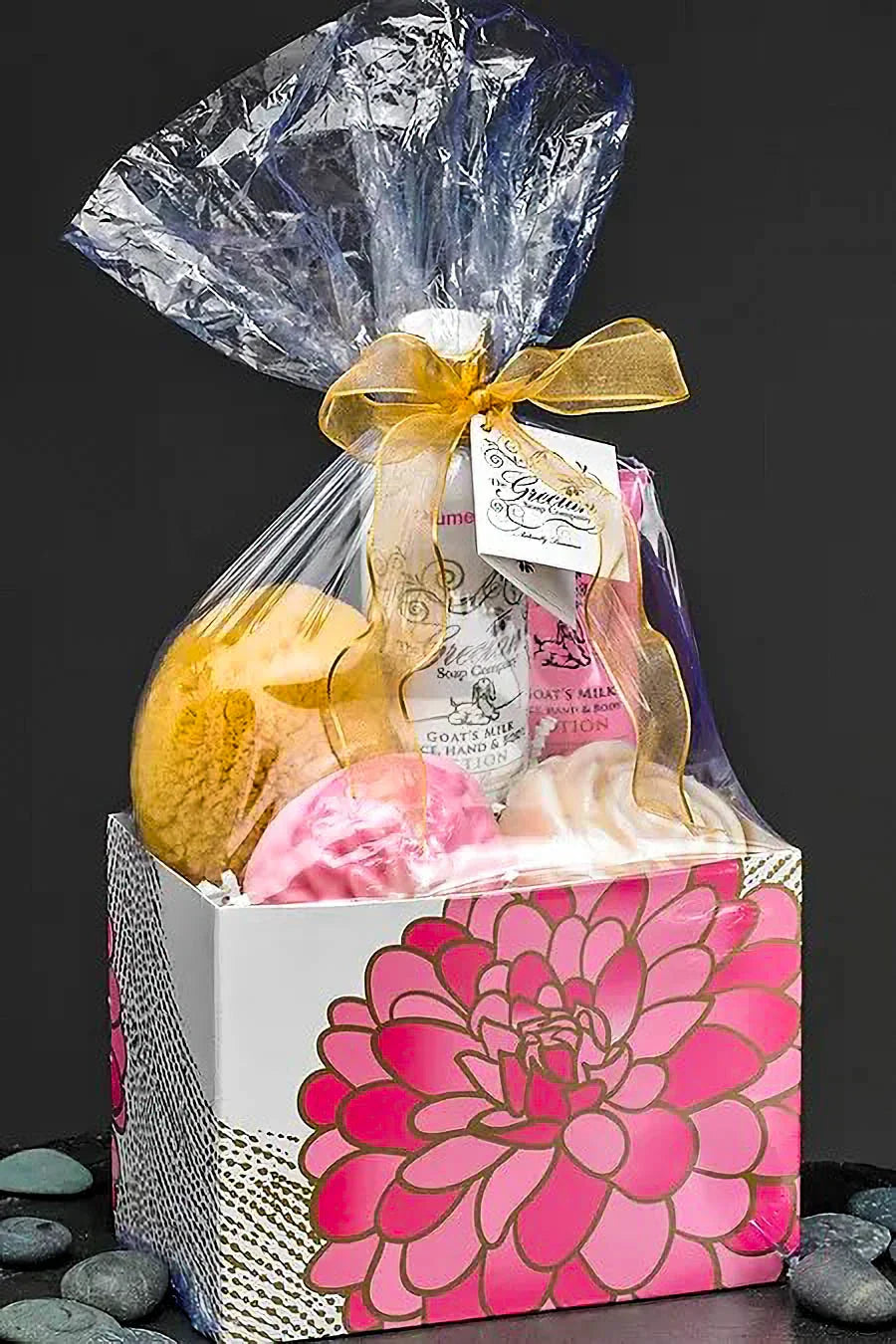 Gift basket with goat's milk lotion and bath items in pink floral box wrapped in clear plastic with gold ribbon