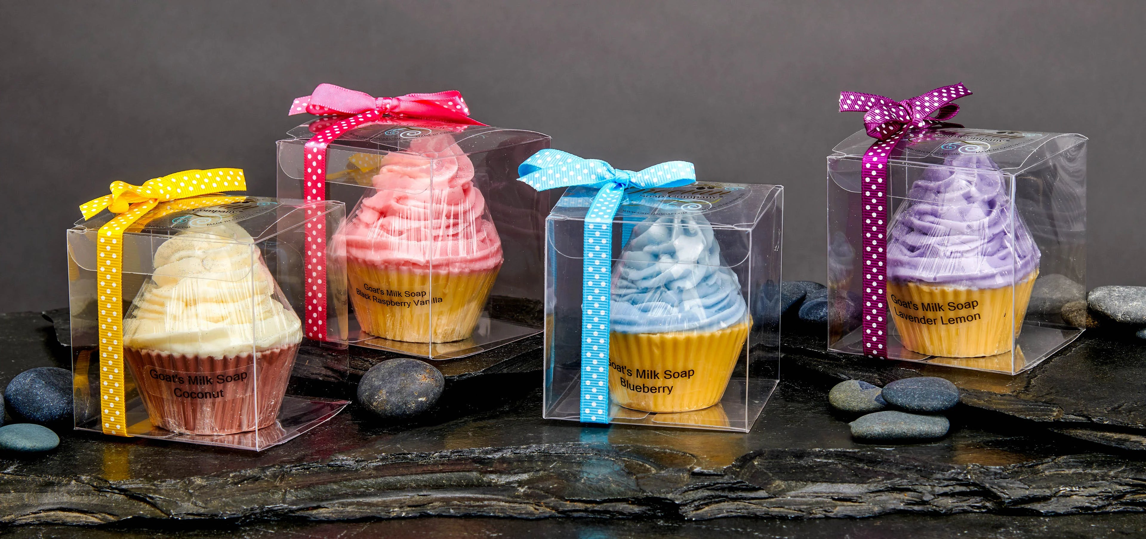 Colorful goat's milk soap cupcakes in clear boxes with ribbons on dark stone and pebbles