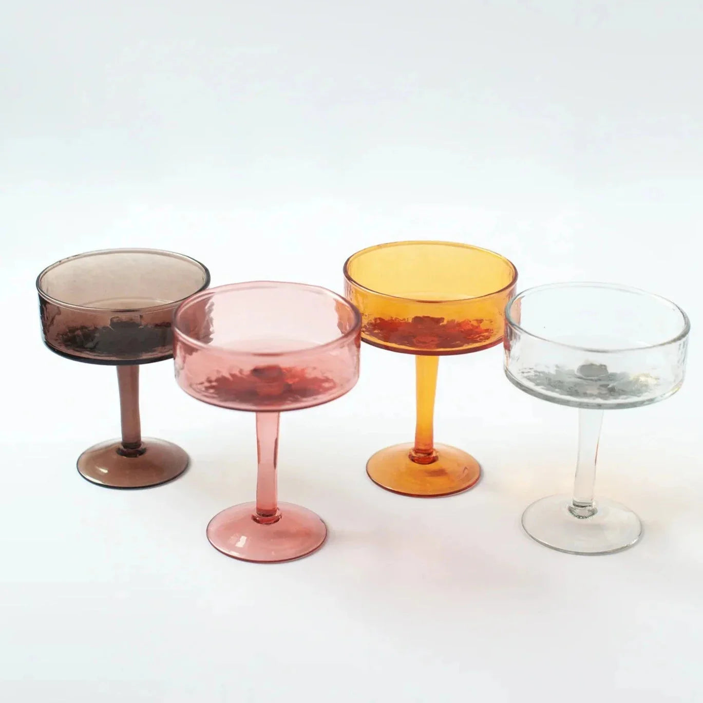 Set of four colorful coupe glasses in brown, pink, amber, and clear on white background