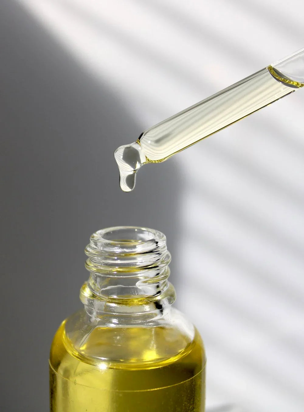 Close-up of glass dropper with golden oil above a small clear bottle, sunlight stripes in background