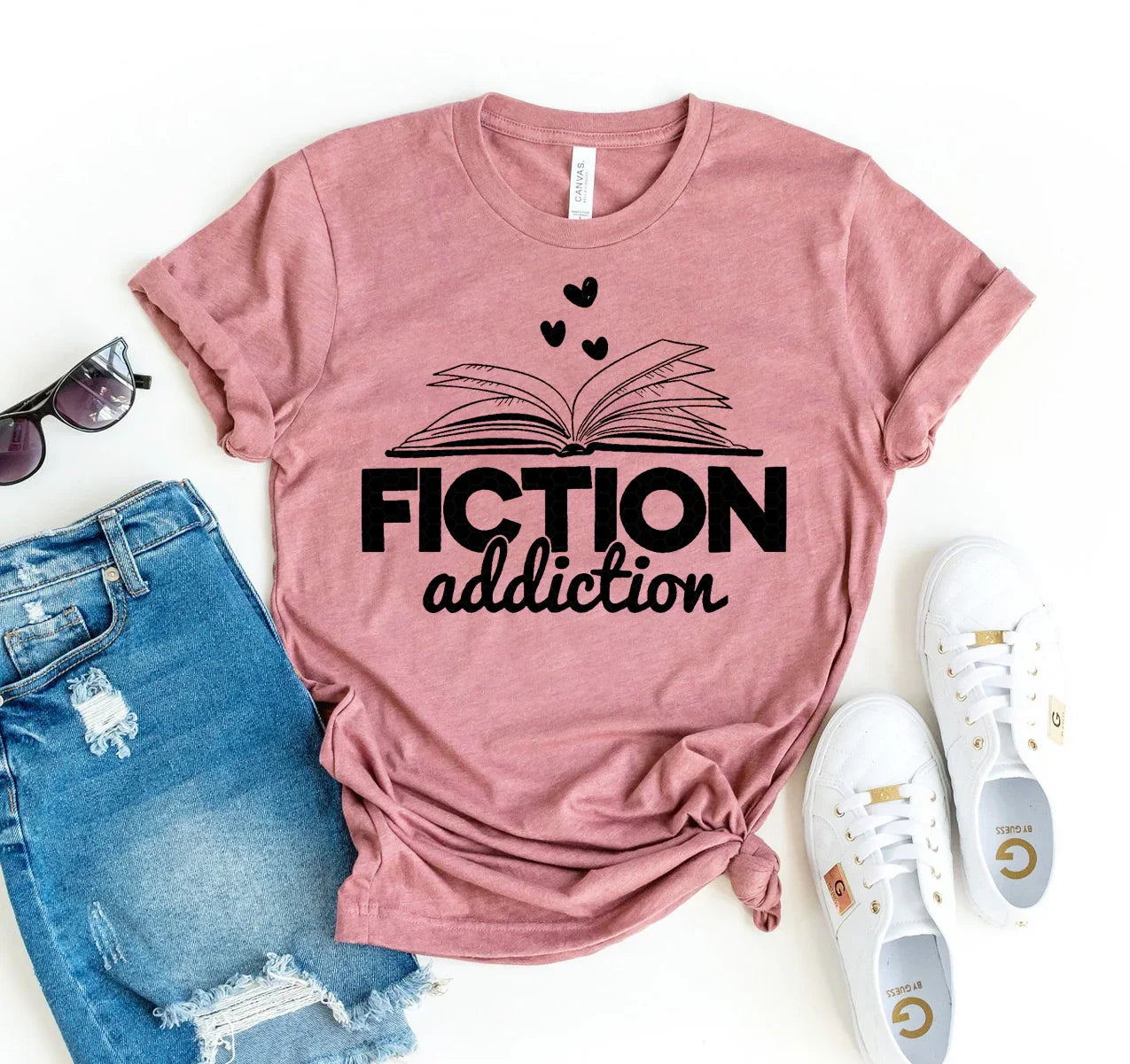Pink t-shirt with black print of open book and hearts reading Fiction addiction, styled with ripped blue jeans, sunglasses, and white sneakers