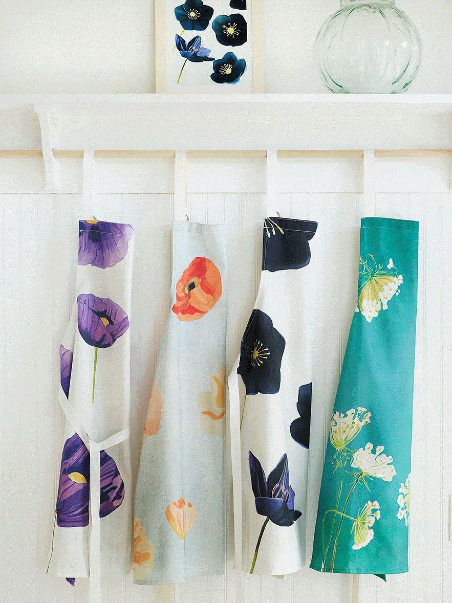 Four floral aprons hanging on white wooden hooks with a floral art print and glass vase above