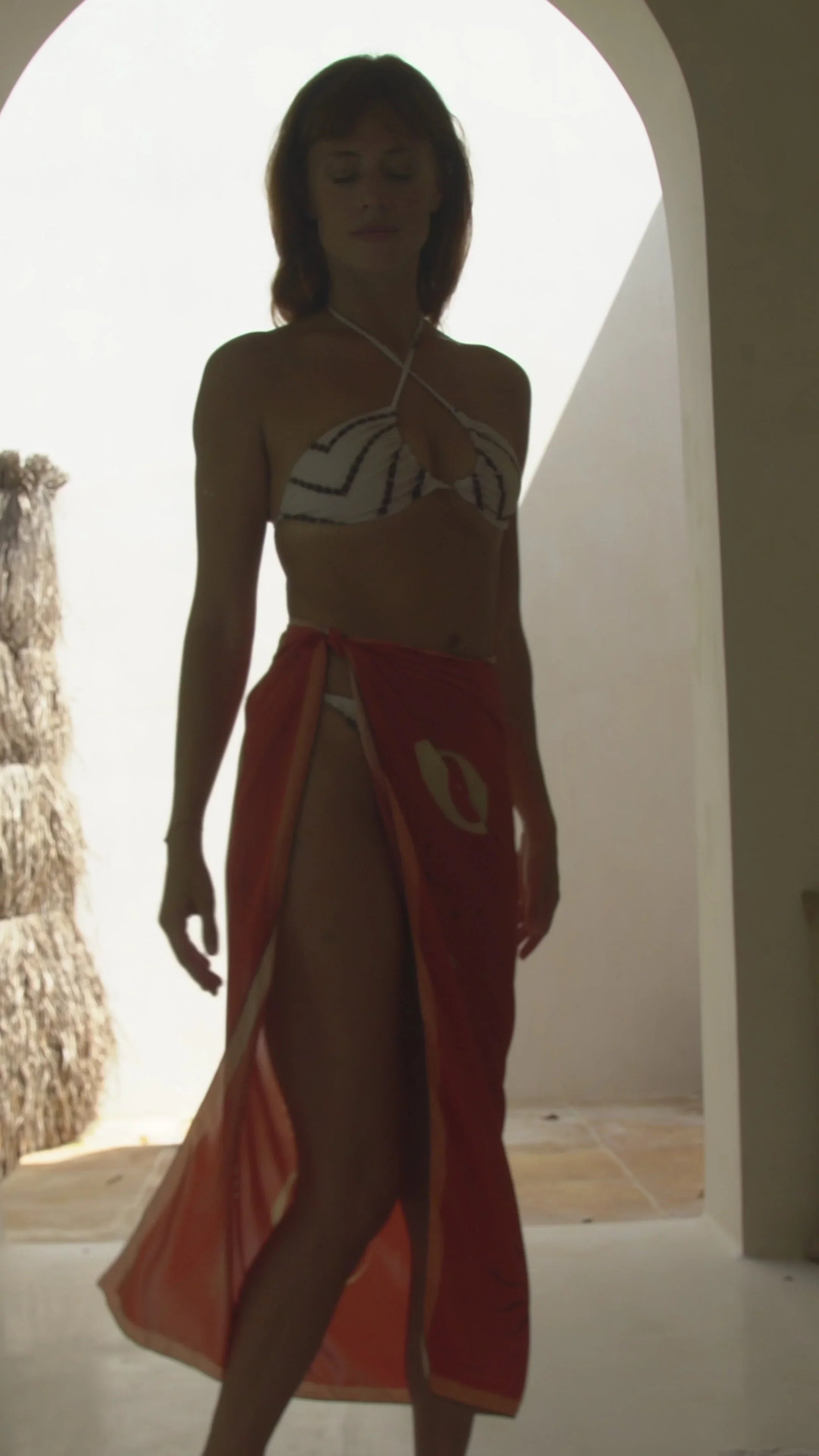 Woman in chevron bikini top and red sarong standing in sunlit archway with tropical decor