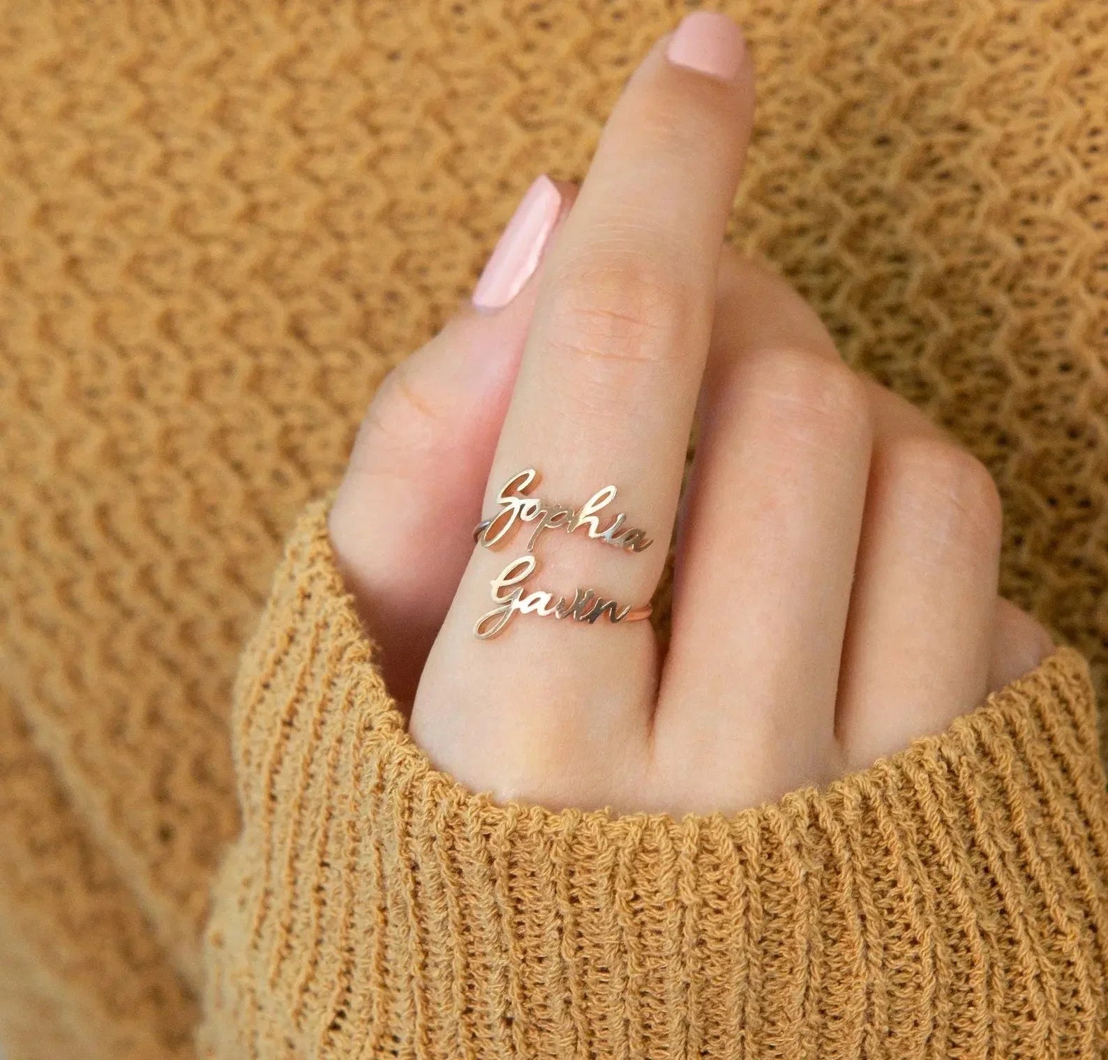 Gold double name ring reading Sophia and Gavin on a woman's finger with pink nail polish and brown knit sweater