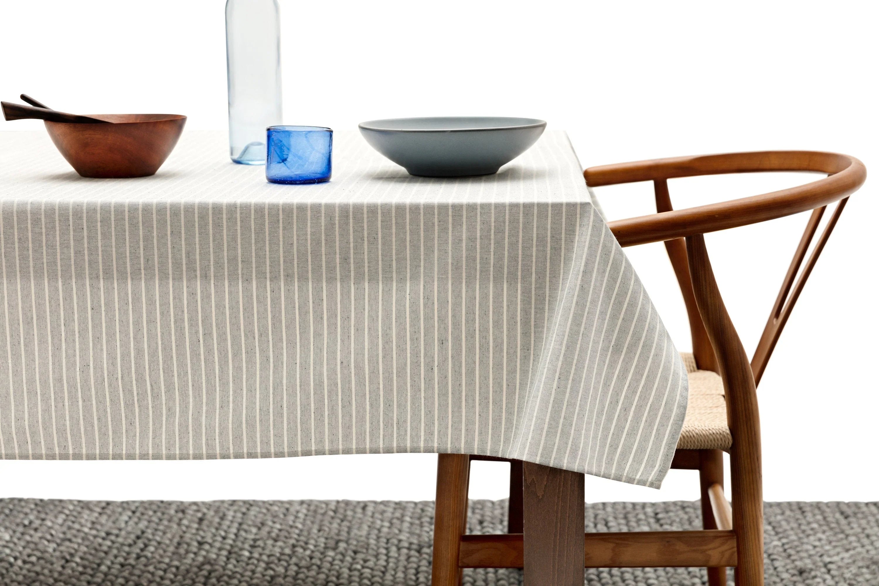 Minimalist dining setup with striped tablecloth, wooden bowl, blue glass, gray bowl, and modern wood chair