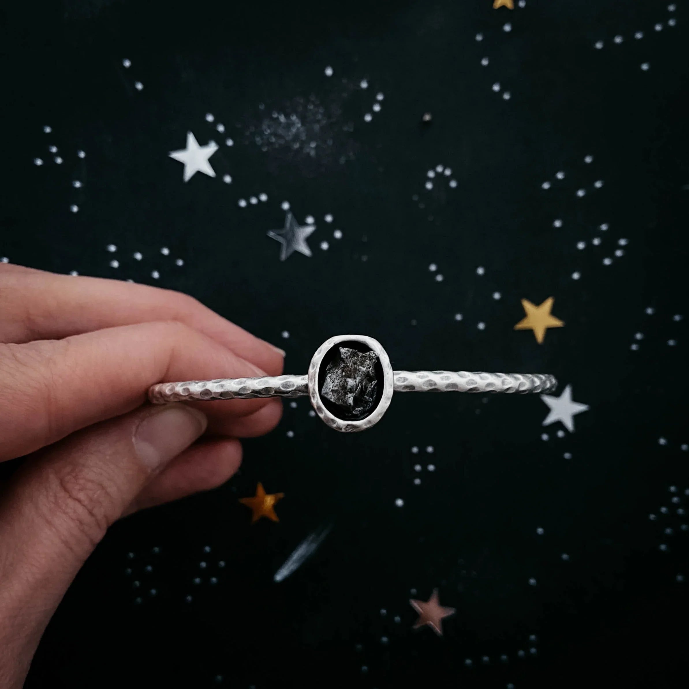 Hand holding a silver hammered bracelet with a rough stone, star background, Qu!rk Boutique