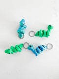 Colorful baby dinosaur keychains in blue and green on a white background, Qu!rk Boutique gift.