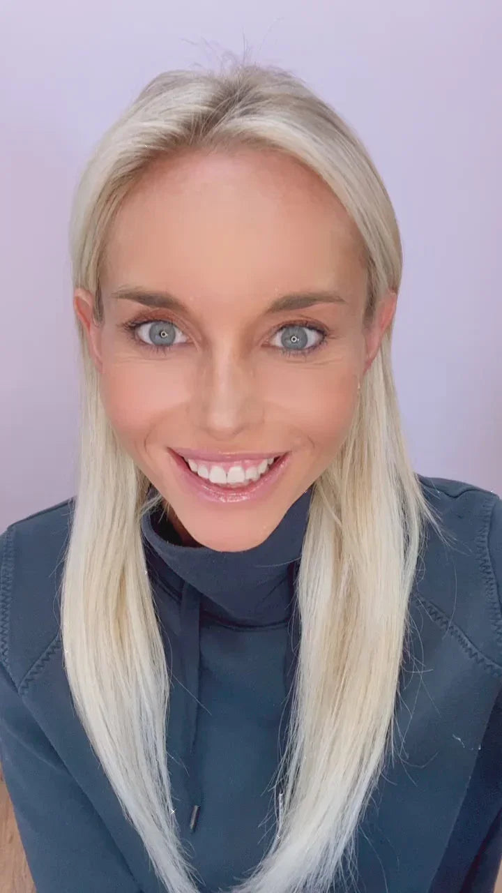 Smiling woman with long blonde hair and blue eyes wearing a dark hoodie, light purple background