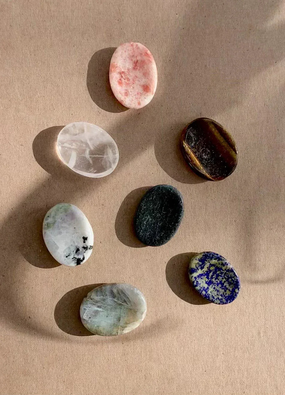 Seven polished oval healing stones of different colors arranged on a beige surface with shadows