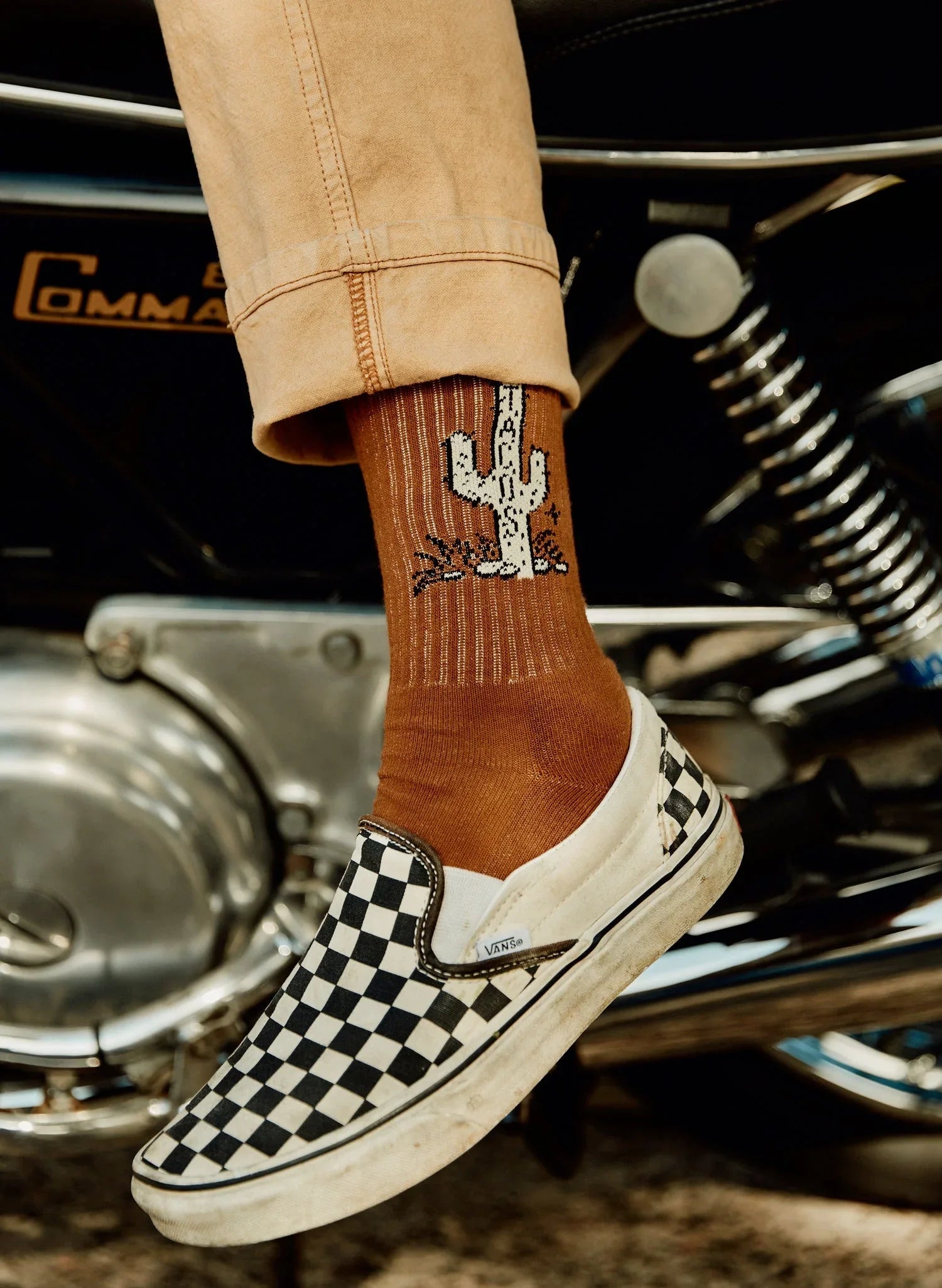 Close-up of brown socks with white cactus design worn with black-and-white checkered Vans slip-on shoes on a motorcycle