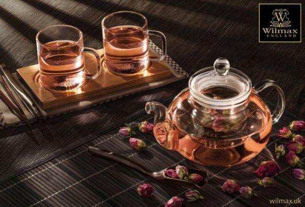 Glass teapot and cups with rose tea on bamboo mat, featuring dried roses and cozy setting